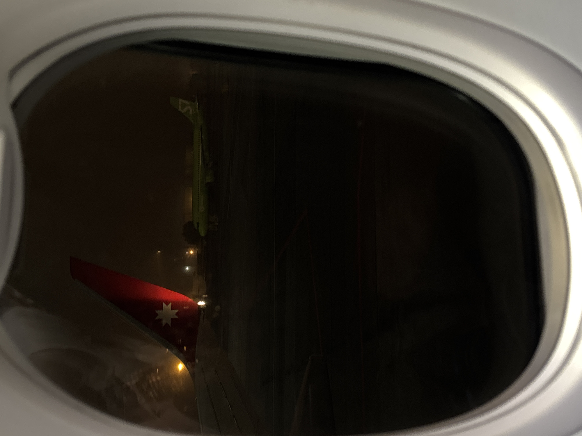 The image shows a view from an airplane window at Domodedovo International Airport in Russia. A red tail fin with the Australian flag design is visible outside the window. In the background, there is a green service vehicle parked near the plane. The scene appears to be at night, with dim lighting present.