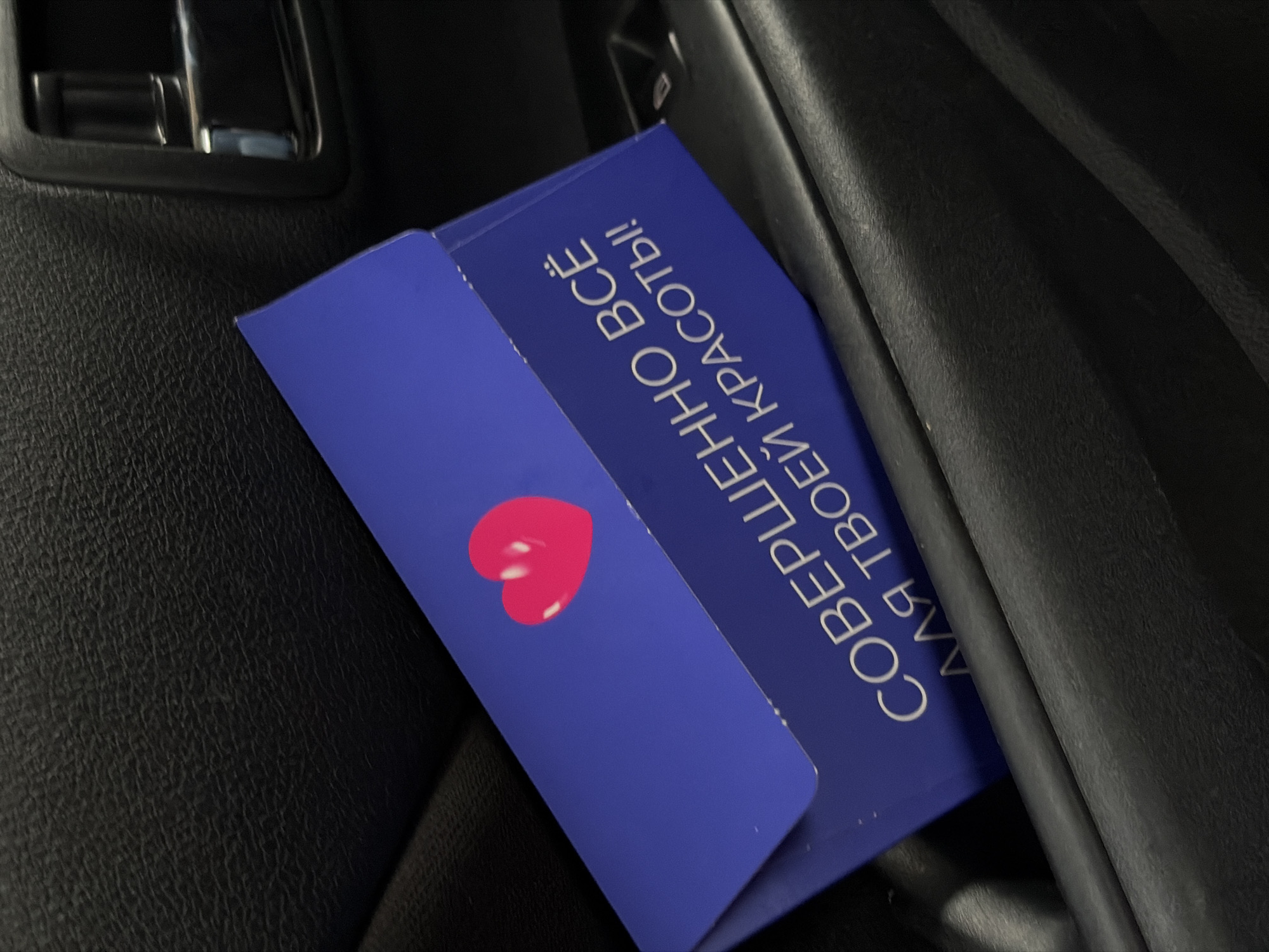 A blue envelope with a pink heart icon is tucked into the dark interior of a car door pocket. White text on the envelope reads "СОВЕРШЕННО ВСЁ ДЛЯ ТВОЕЙ КРАСОТЫ!" which translates to "Absolutely everything for your beauty!". The car's interior appears to be made of black textured material. The image is cropped to focus on the envelope and the immediate surrounding car interior. The scene is set in Zubovka, Russia.