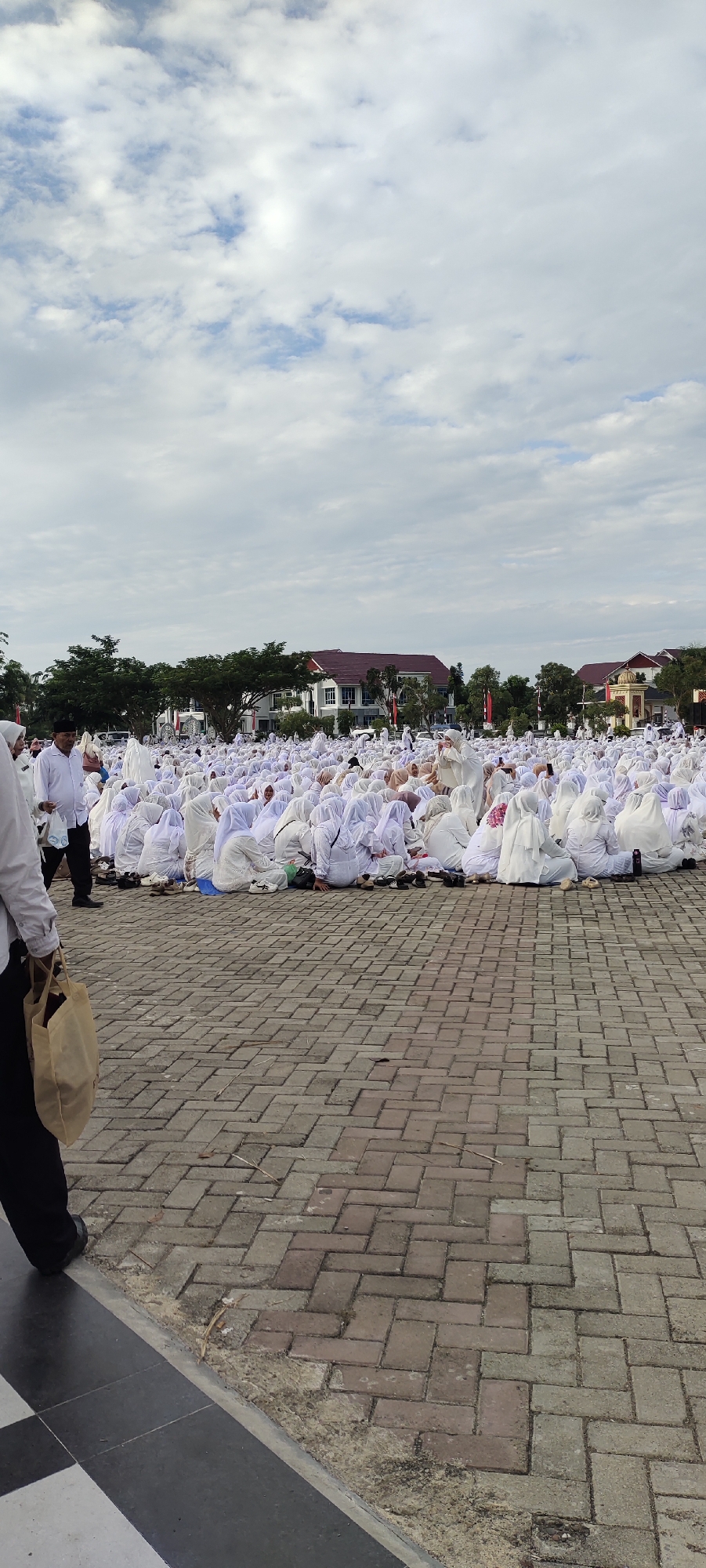 The image depicts a large gathering of people, predominantly women, dressed in white attire, seated on a brick-paved outdoor area. The gathering appears to be taking place in an open field or square, with buildings and trees visible in the background. The sky is overcast with scattered clouds. A few individuals in the foreground are partially visible, including a person carrying a beige tote bag and another man in a white shirt. The overall scene suggests a communal event or religious gathering. The location is identified as Meunasahaluedrienlhoksukon Barat, Indonesia.