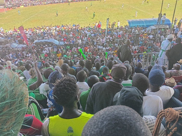 This media file captures a vibrant scene at an outdoor football (soccer) stadium in Siaya, Kenya, during a match. The primary subjects are a large crowd of spectators filling the stands, along with football players visible on the green pitch.
The image is taken from a raised vantage point among the lower stands, looking over the heads of the immediate crowd towards the field. On the lush green pitch, several players in light-colored uniforms are actively engaged in the game. A blue dugout or shelter can be seen on the far side of the field, beneath another section of spectator seating.
The audience is densely packed, with many individuals wearing green clothing, suggesting support for a particular team. The atmosphere appears lively, with several spectators raising their hands, and green vuvuzelas are visible among the crowd. A prominent red flag is also seen waving in the mid-ground. Metal barriers separate different sections of the stadium. A dark-colored vehicle is partially visible above a section of the crowd, possibly a security or broadcast vehicle.
The bright lighting indicates it is daytime with sunny weather. Visible text on various shirts worn by spectators includes "TAPES," "H.F.," "BLOO," and "R.F.E." A yellow vest in the foreground features a circular logo, but the text within it is not clearly legible. The overall impression is that of an enthusiastic and well-attended local sports event in Kenya.