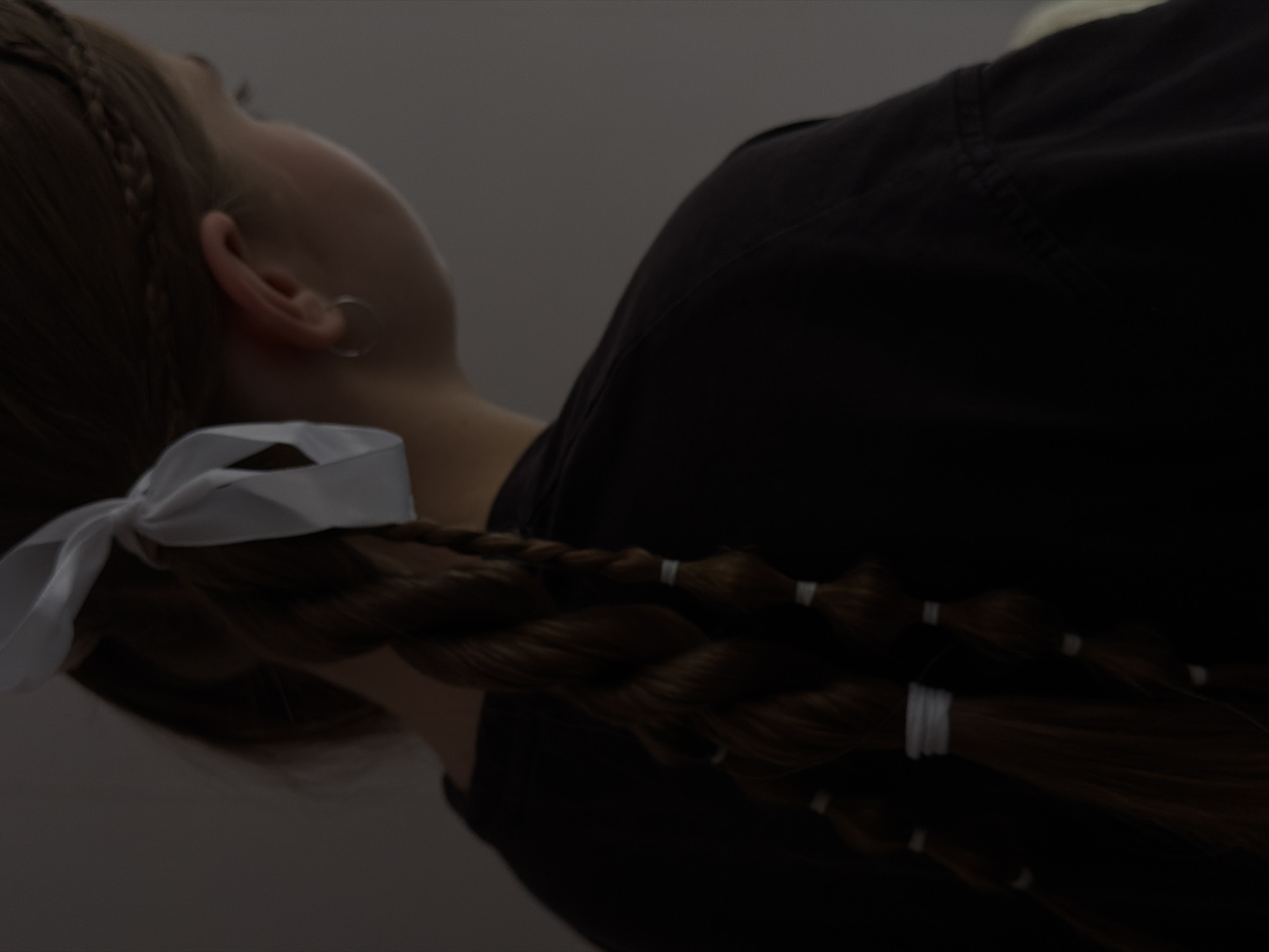 The image shows a close-up, angled view of a person, likely female, from the chin and ear down to the back. The person has long, dark brown hair styled in a thick braid. A prominent white ribbon is tied into a bow at the top of the braid, near the neck. Further down the braid, several smaller white elastic bands or ties segment the hair. A silver hoop earring is visible in the person's left ear. The individual is wearing a dark, possibly black, garment, only a portion of which is visible on the shoulder and back. The person's head is tilted upwards, suggesting they might be lying down or looking up. The background is a plain, dimly lit, grayish surface, indicating an indoor setting. No specific activity is taking place, and no text is visible. The overall mood is subdued due to the low lighting. The location context is Perm, Russia.
