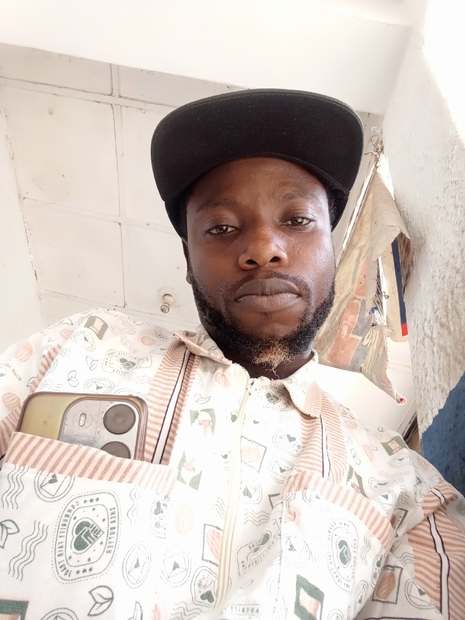 A medium shot captures a man from the chest up. He wears a black flat-brimmed cap and a patterned, light-colored shirt with a zipper closure. A smartphone is visible in the chest pocket of his shirt. The man has dark hair and a beard with some lighter strands. The background consists of a white, tiled ceiling and a textured white wall. Part of a fabric item with red and blue stripes hangs beside the wall. The location is Unknown, Unknown.