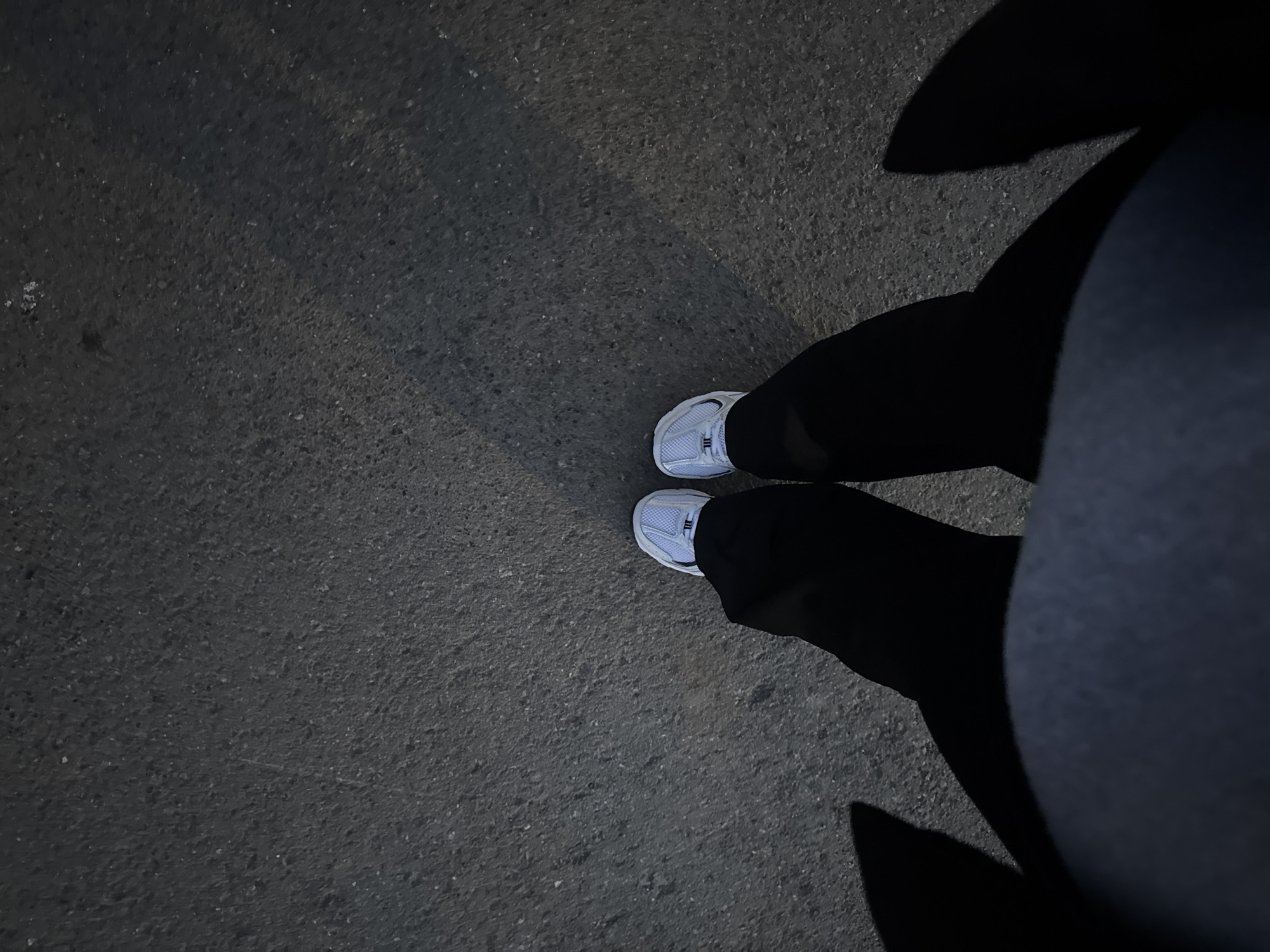 The image presents a top-down perspective of a person's feet and lower legs, illuminated against a dark, granular ground surface, likely asphalt or concrete. The person is wearing white sneakers with visible laces and dark trousers. The upper parts of the legs and body are mostly obscured by large, dark shadows cast dramatically across the right side of the frame, suggesting a strong, localized light source, likely from above and to the left of the scene. The shadows create abstract, elongated shapes on the ground.
The scene appears to be set at nighttime or in very low light conditions, with artificial illumination highlighting the texture of the ground and the white sneakers. The ground itself shows variations in tone, indicating subtle differences in texture and illumination. There are no other discernible objects or individuals in the immediate vicinity.
The activity depicted is simply the person standing or possibly in mid-stride. The overall mood is quiet and still, emphasizing the interaction of light and shadow on the urban ground. The location context provided indicates this scene is in Perm, Russia. No visible text is present in the image.