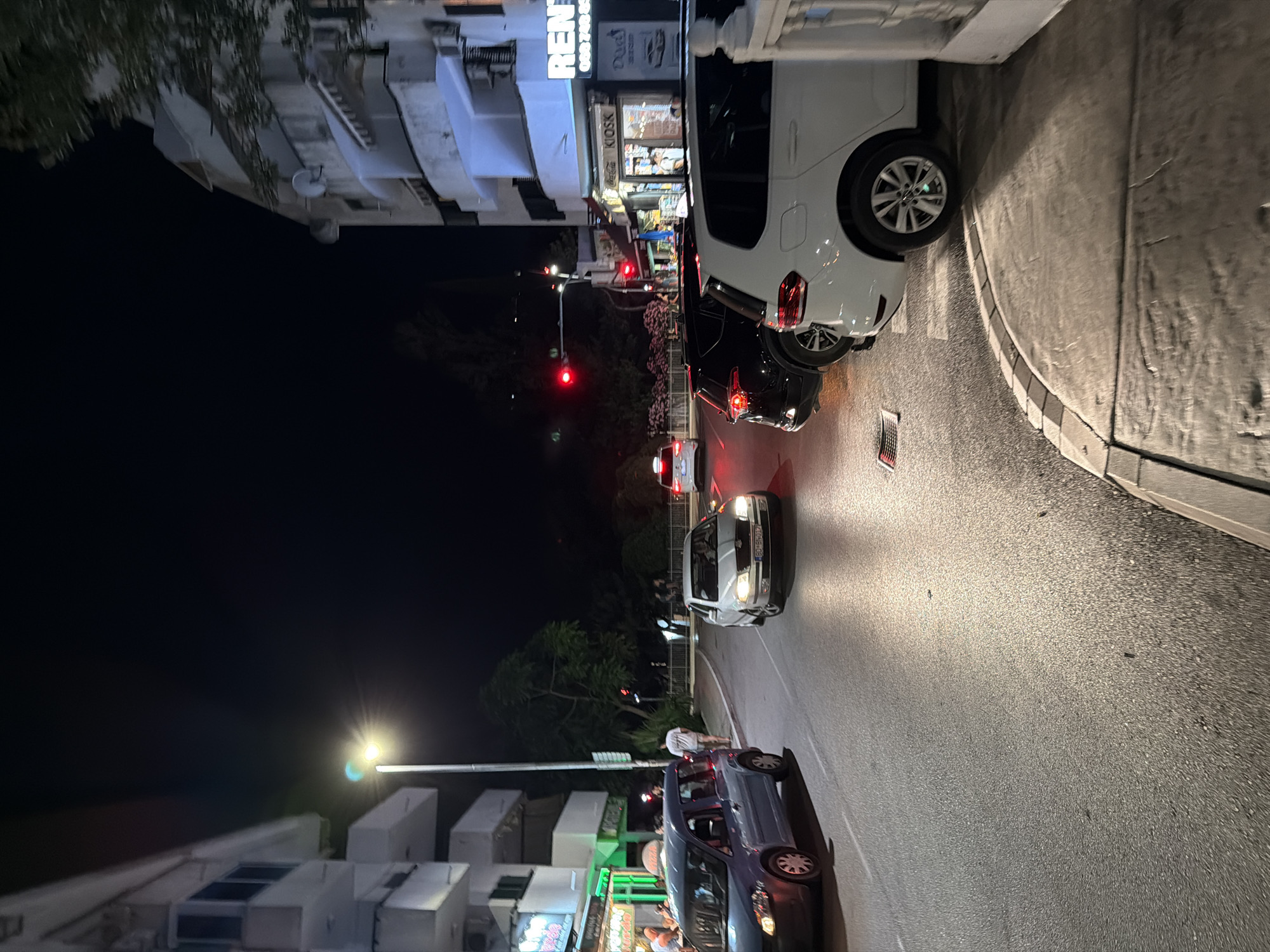 The image depicts a street scene at night in Budva, Montenegro. The street is paved and appears wet, reflecting the surrounding lights. Several cars are present, including a blue minivan on the left, a silver sedan in the middle, a black SUV behind the sedan, and a white SUV on the right. Pedestrians are visible, with one person in a white t-shirt standing near the blue minivan and others further down the street. Buildings line the right side of the street, featuring illuminated signage, including one that reads "RENT." Traffic lights are visible, with at least one showing red. Trees and foliage are present in the background, obscuring parts of the sky. The overall atmosphere suggests a bustling urban environment at night.