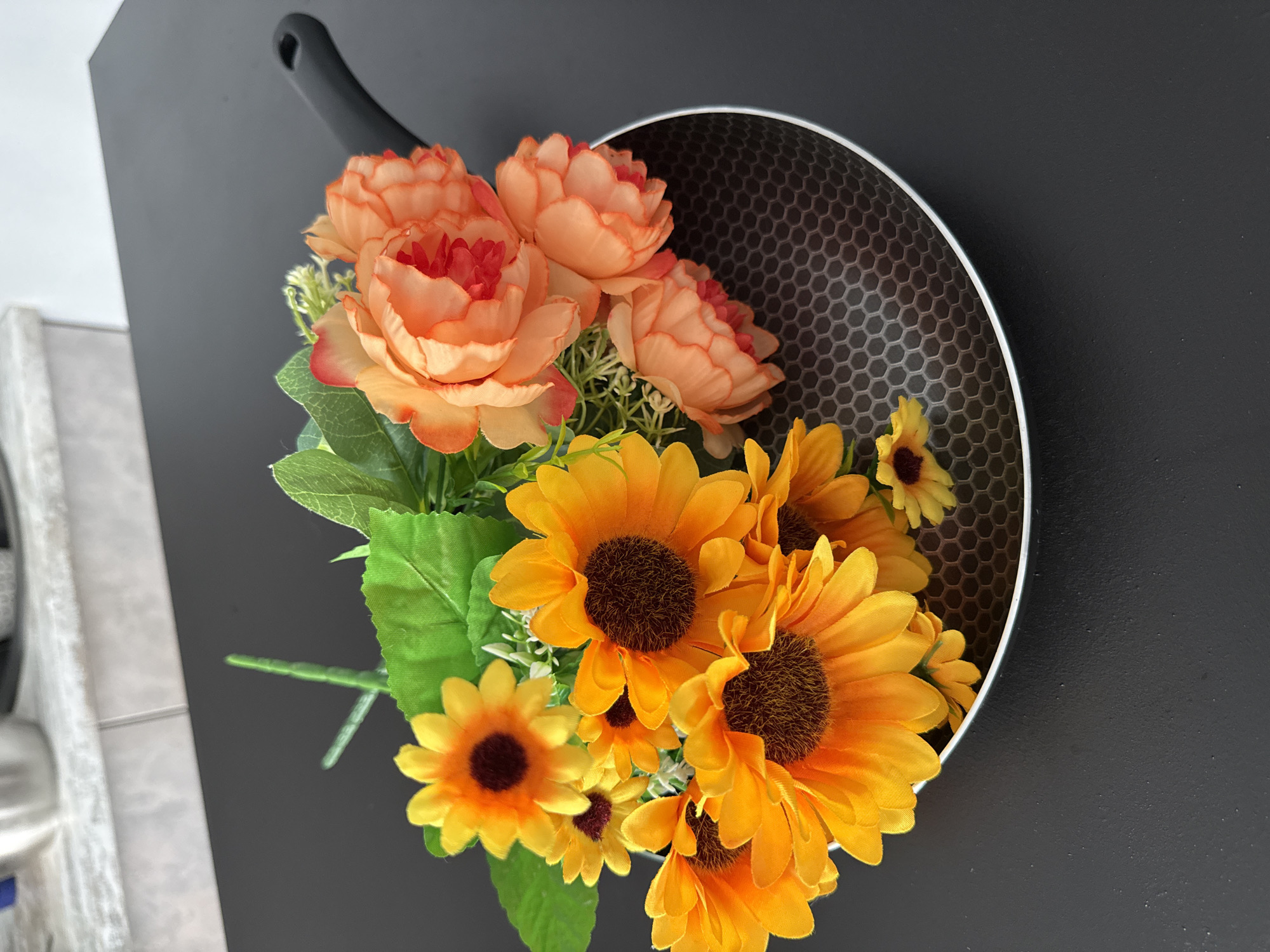A top-down shot displays a black frying pan filled with artificial flowers. The flowers consist of orange-peach peony-like blooms and yellow sunflowers, interspersed with green leaves and stems. The frying pan, equipped with a black handle, features a patterned surface on the inside. The scene is set on a black surface. The location is not provided.