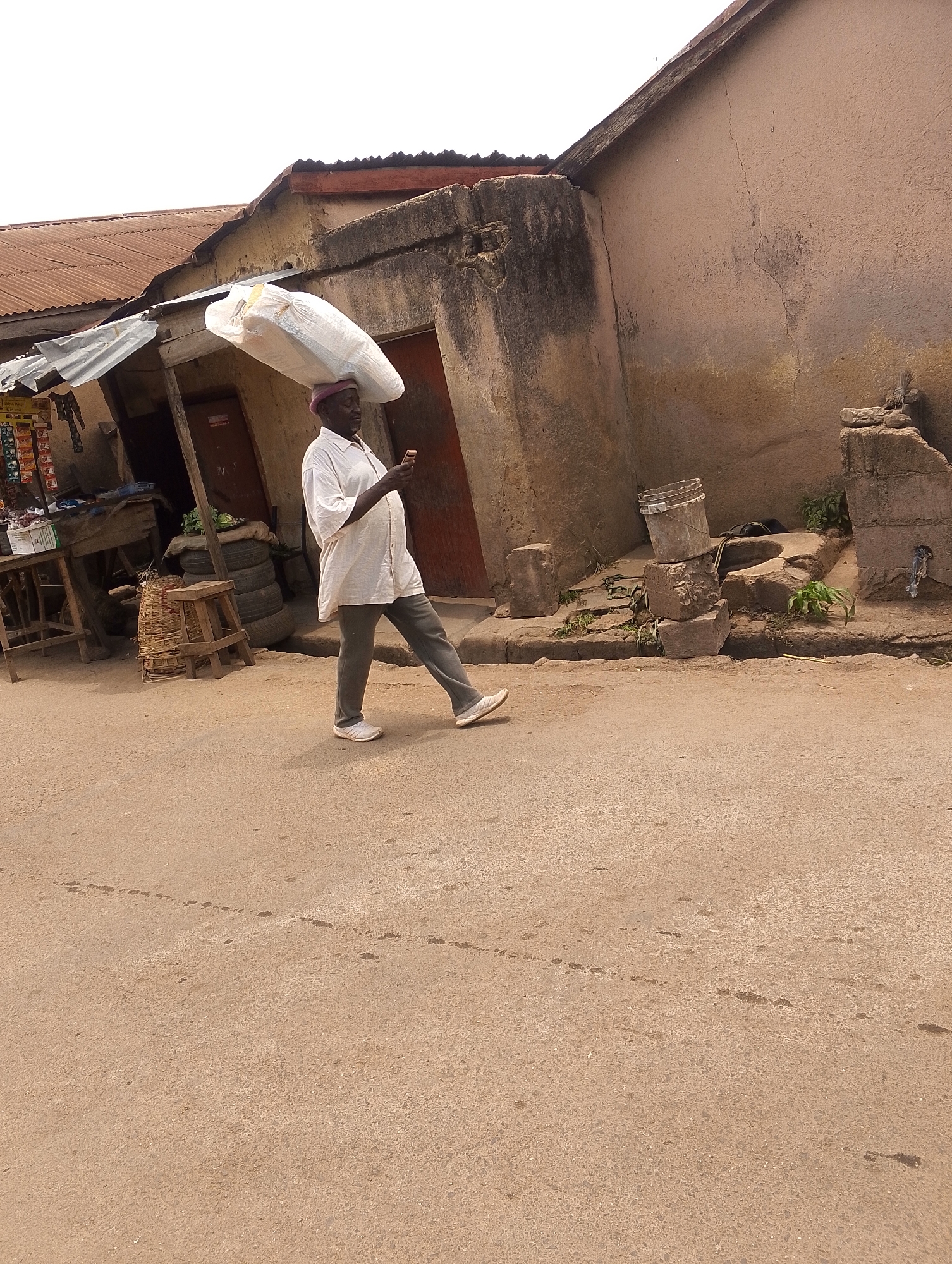 The image depicts an elderly man walking on a street in Jos, Nigeria. He is carrying a large, white, plastic-wrapped load on his head and using a cell phone. He is wearing a white, short-sleeved shirt, gray pants, and white sneakers, along with a purplish cap.
The street appears to be a local road. To his left is a small, dilapidated shop with various items displayed. Behind him are buildings with weathered walls and corrugated iron roofs. There are also stacked tires and a makeshift outdoor seating area near the shop. The street itself is unpaved and appears sandy or dusty.
