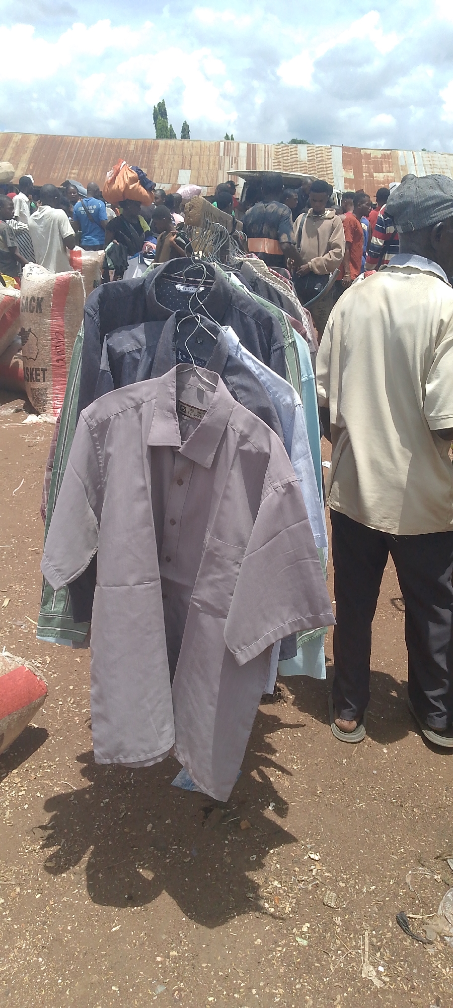 The image depicts an outdoor market scene in Tor-Donga, Nigeria, under a cloudy sky. In the foreground, a rack of men's shirts is displayed. The shirts are hung on hangers and appear to be made of cotton or a similar fabric, with various colors including light gray, navy blue, light blue, and a striped green and white shirt. One light gray shirt in the front is a short-sleeved button-down. Behind the shirts, a large burlap sack labeled "SACK BASKET" is visible, with part of its contents or branding showing.

In the background, a crowd of people is milling around, suggesting a busy marketplace. Many individuals are dressed in casual clothing. Some people are carrying bundles or bags. A corrugated metal structure, possibly a building or stall roof, runs across the upper portion of the background. There are trees visible in the distance. The ground is composed of dirt and small debris. A person wearing a light-colored short-sleeved shirt and dark pants, with sandals, stands to the right of the clothing rack, partially obscuring the background crowd. The overall impression is of a bustling local market environment.
