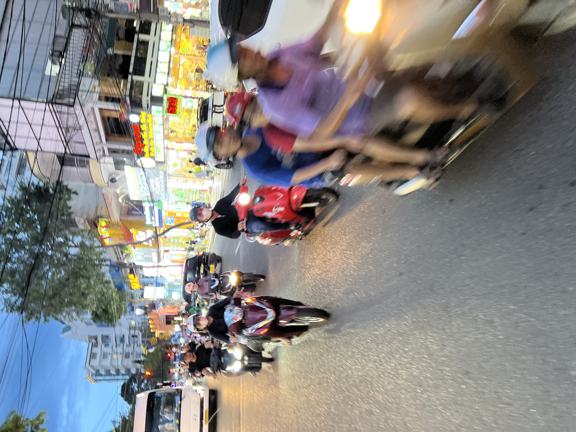 The image depicts a street scene in Nha Trang, Vietnam, likely at dusk or early evening, as indicated by the twilight sky and illuminated buildings. The foreground is dominated by a busy road with several individuals on motorbikes, creating a sense of motion blur.
In the immediate foreground, a person wearing a purple shirt and dark shorts is riding a motorbike, with their right hand visible and illuminated by the motorbike's headlight, suggesting they are actively driving. Behind them, another person in a blue shirt and dark shorts is riding on the same motorbike. Further ahead, a person in a red shirt is also on a motorbike.
To the left, a burgundy motorbike is being ridden by someone wearing a black shirt and a helmet. Another motorbike with a person in a dark shirt and helmet is slightly behind and to the left. A white van is visible on the far left of the image.
The background shows a bustling urban environment with multi-story buildings lining the street. Many of these buildings feature illuminated signage and windows, suggesting commercial establishments. Trees are interspersed along the street. The overall impression is a lively and active street in an Asian city.