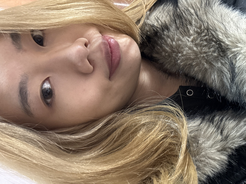 The image displays a close-up, selfie-style portrait of an individual, an adult female of East Asian descent, located in Shapingba District, China. The subject possesses blonde-dyed, wavy hair, and fair skin. Her eyes are dark, appearing to wear light-colored contact lenses. Her eyebrows are natural, and her lips, colored light pink, form a subtle smile with the mouth slightly parted. She is wearing a dark garment, identified as a jacket or coat, featuring a prominent faux fur collar in shades of grey and white/light brown. Two silver-colored snap fasteners are visible on the black placket of the garment. The individual gazes directly into the camera. The background is indistinct and light-colored, suggesting an indoor environment with soft, diffuse illumination.