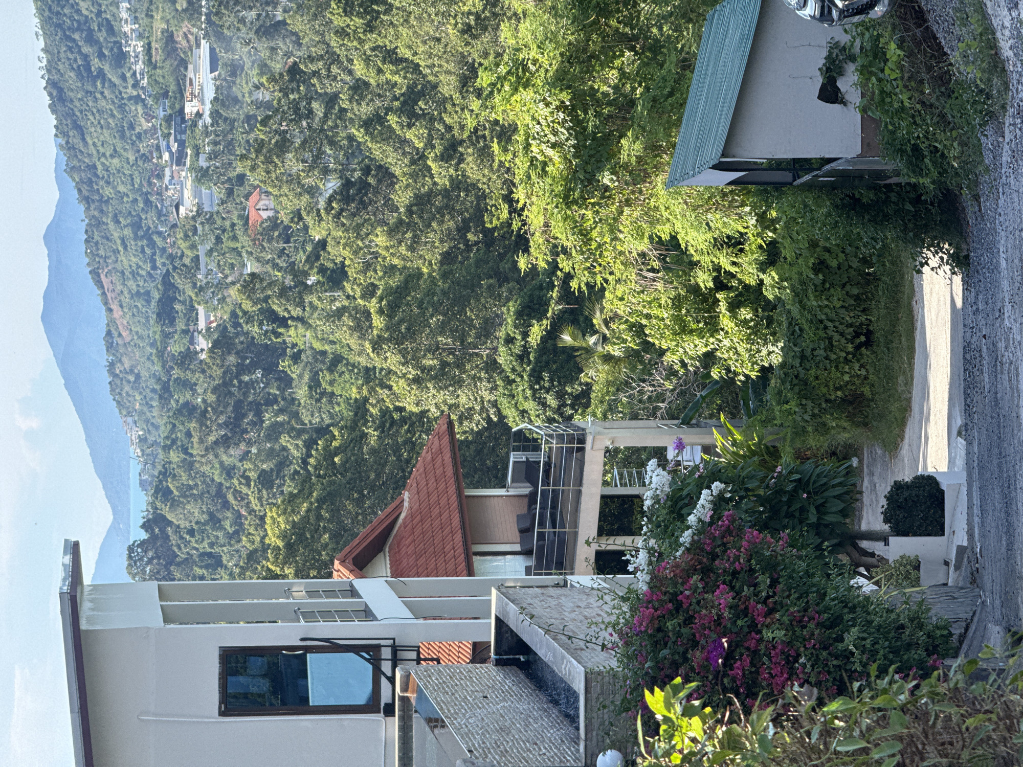In Baan Chaweng, Thailand, a view overlooks a lush, verdant landscape dotted with residential buildings nestled among dense foliage. A paved driveway slopes downwards from the foreground, bordered by gray gravel on the right. To the left, a modern multi-story building features a clean white facade with a dark-trimmed window and a balcony with glass railings. A water feature with stacked stone accents is visible on the upper level of this building. Below, vibrant pink and white bougainvillea flowers cascade down, partially obscuring a lower section of the building. A second, smaller building with a red tiled roof and a balcony furnished with dark outdoor seating is situated further down and to the right. Beyond these structures, the landscape transforms into a rolling hillside covered in a thick canopy of various green trees, stretching towards distant mountains under a bright, clear sky. On the far right, a small, grey shed-like structure with a green corrugated metal roof is partially visible, with a portion of a car in the extreme right edge of the frame. The scene is bathed in sunlight, casting shadows and highlighting the rich textures of the vegetation and architecture.