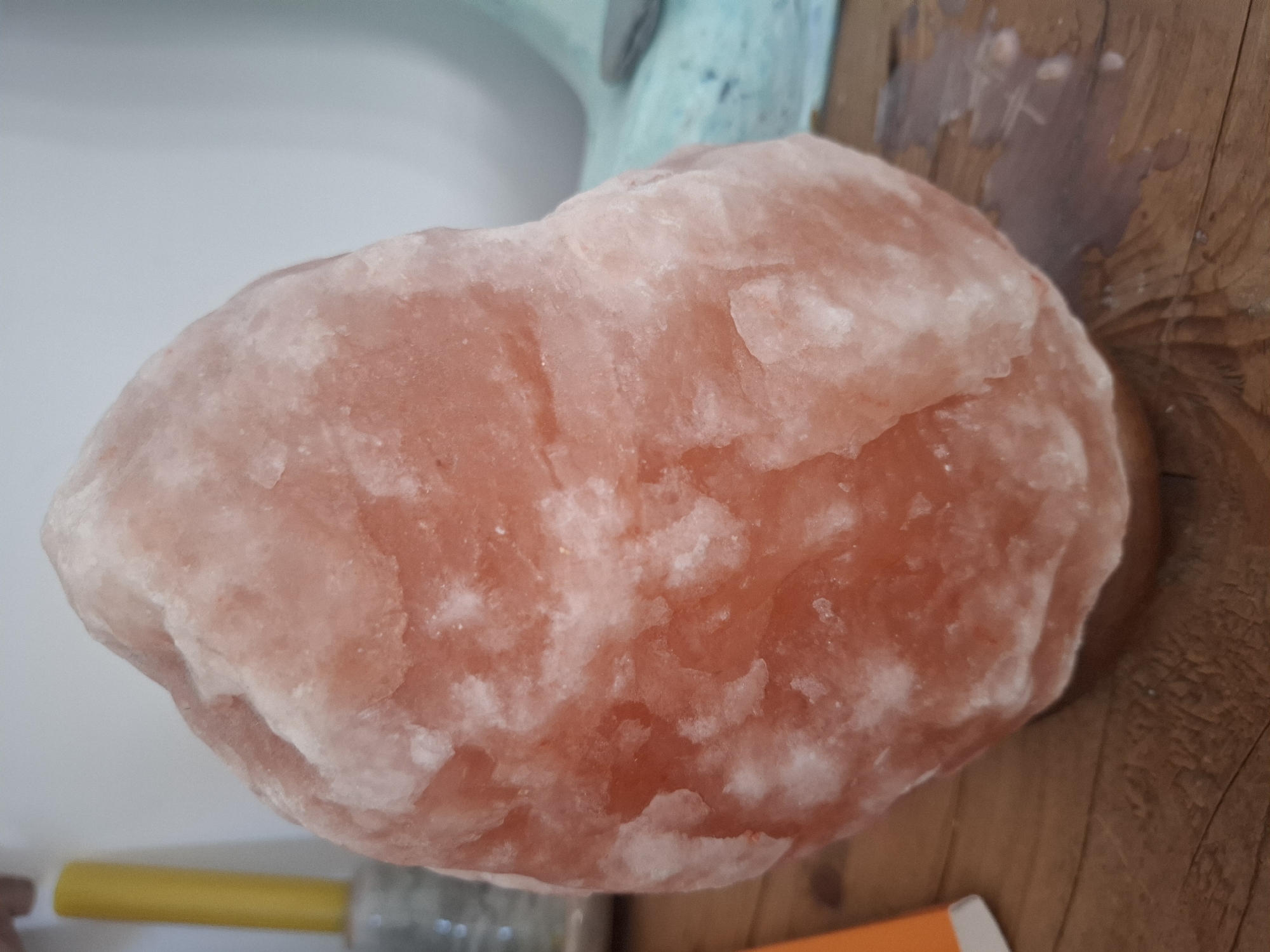 A Himalayan salt lamp sits on a wooden surface. The lamp is roughly egg-shaped and appears to be an unprocessed chunk of pinkish-orange salt with white crystalline deposits. In the background, to the left, a yellow cylindrical object is visible, possibly a candle or a dispenser. To the right of the lamp, a blurry, light blue object with some texture can be seen. The wooden surface shows grain patterns and a few water spots. The background wall is a light, neutral color. No people or actions are depicted. The image captures a still life arrangement of objects. The location in Montreuil, France, is not evident from the visual information provided.