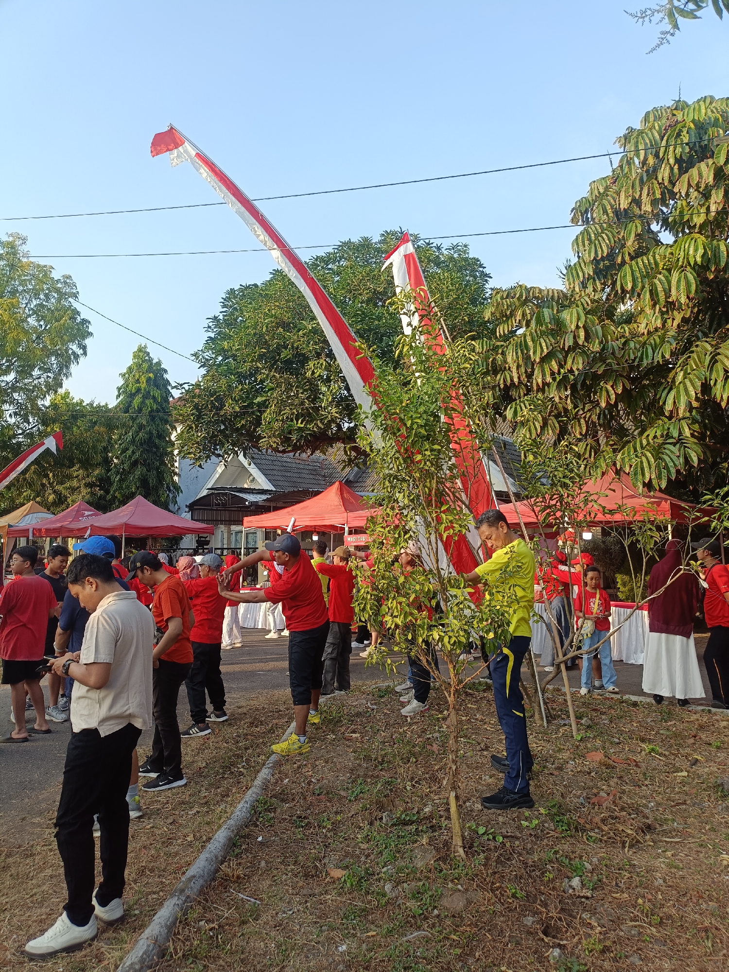 The image captures an outdoor scene, likely in the city of Kediri, Indonesia, with a group of people gathered. In the foreground, several individuals are standing, some wearing red shirts and black pants, others in lighter-colored attire. A man in a light beige polo shirt and black pants is looking at his phone. Another man in a yellow shirt with blue accents and dark pants is standing near a small tree. In the background, multiple red tents are set up, suggesting a market or event. Two large Indonesian flags, red and white, are prominently displayed, attached to tall poles that extend into the sky. The flags appear to be part of a celebratory or ceremonial gathering. Trees and foliage are visible, along with a building in the distance. The ground is covered with dry grass and dirt, with a fallen log in the lower left portion of the image. The overall atmosphere suggests a daytime event under a clear sky.