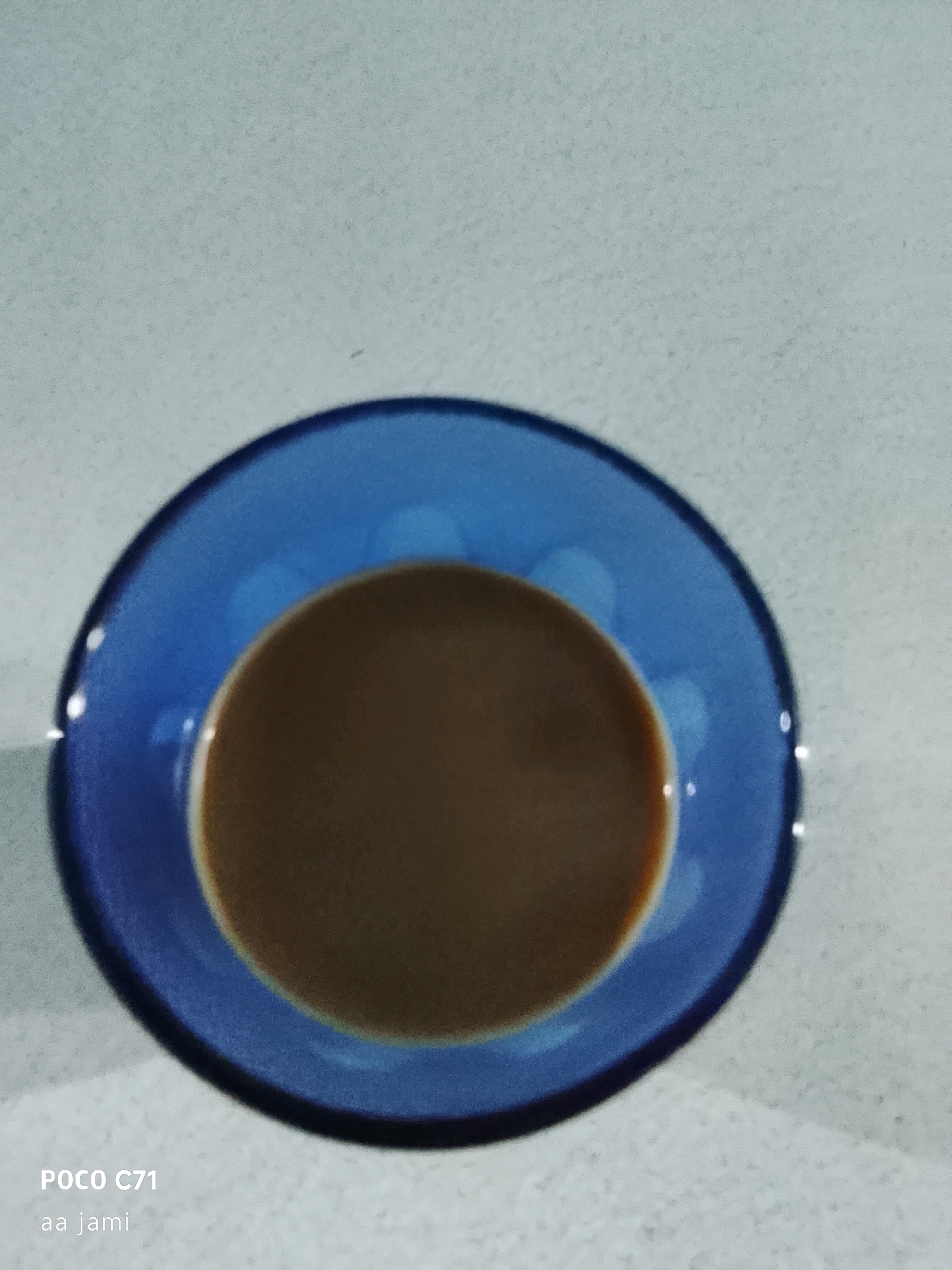 A close-up, overhead shot captures a cup of coffee on a textured, light-colored surface. The cup, a vibrant blue, has a wide rim and is filled with a light brown liquid, likely coffee. The surface beneath appears to be a speckled countertop or placemat. In the lower left corner, text reads "POCO C71" and "aa jami". The image quality is slightly grainy, suggesting it was taken with a mobile device in low light. There is no indication of the location in Tasikmalaya, Indonesia, in the visual content of the image.