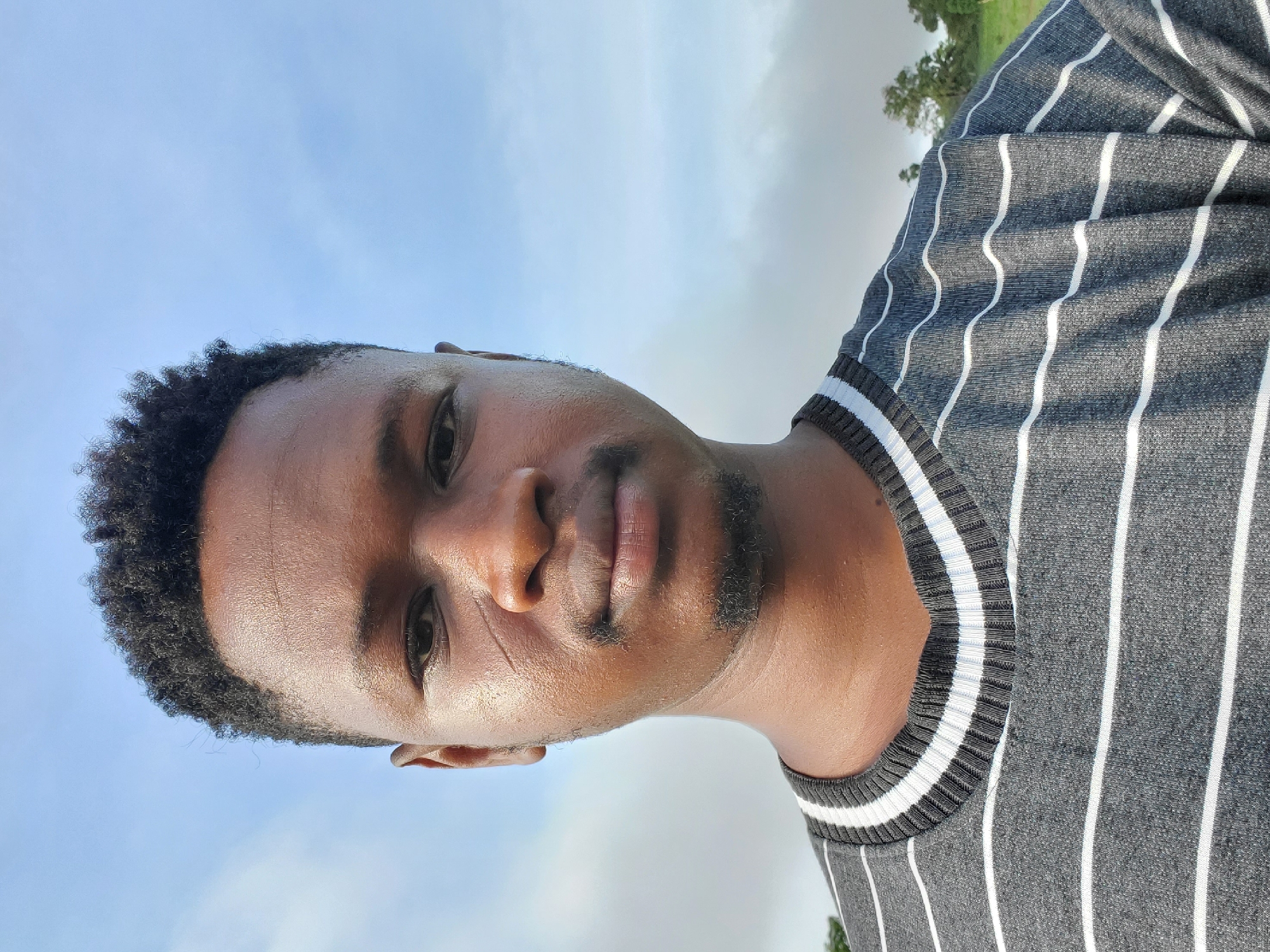 A portrait of a Black male is taken outdoors against a partly cloudy sky. He is wearing a gray and white striped shirt. The location is identifiable as Mararaba, Nigeria, based on the subject's likely place of residence and general environmental features. No specific landmarks or details are present to precisely pinpoint a locality within the city.