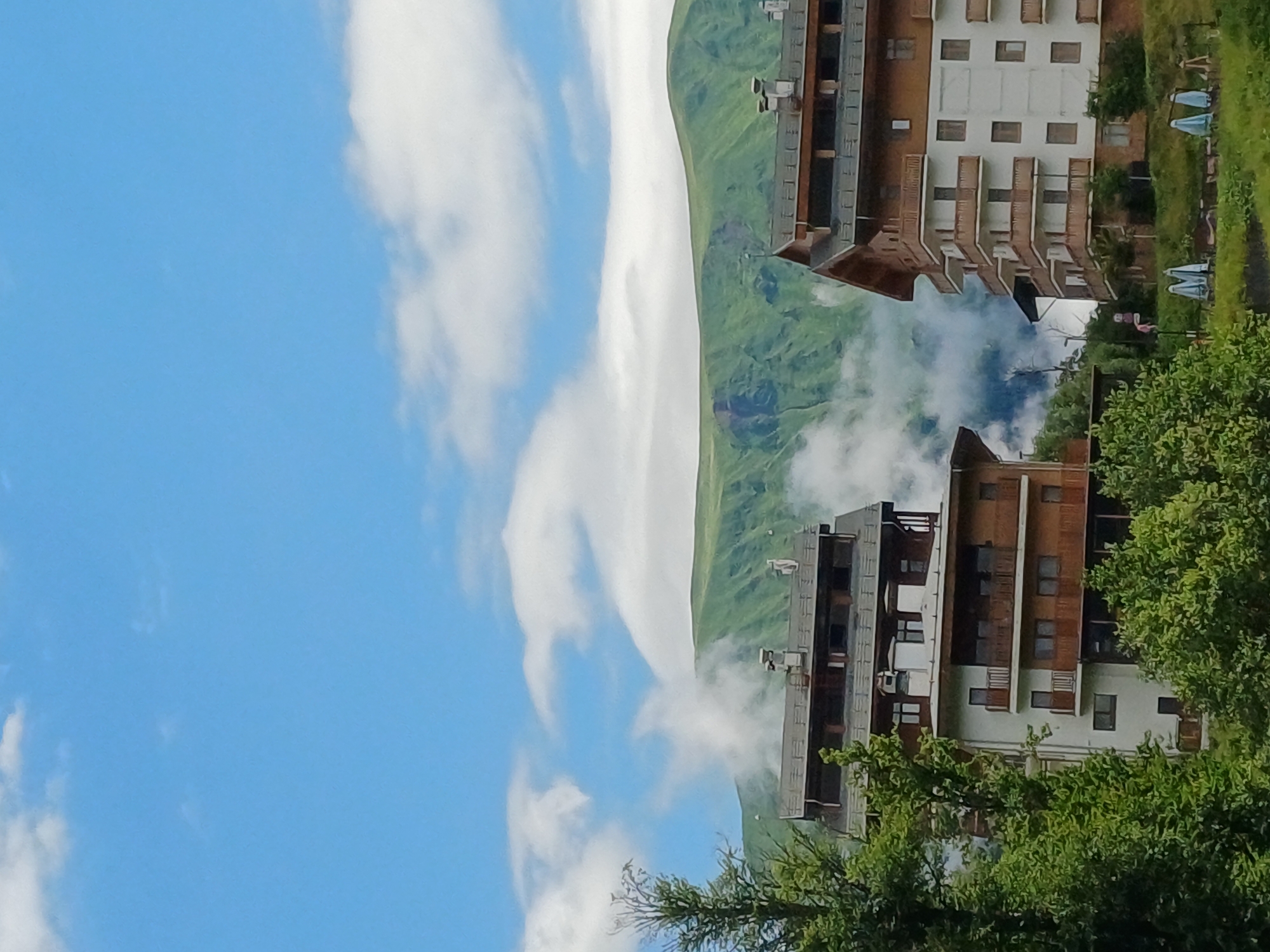 The image captures a scenic view in Estosadok, Russia, featuring modern multi-story buildings nestled against a backdrop of lush green mountains. The sky is a vibrant blue, dotted with white, fluffy clouds, some of which partially obscure the mountain peaks, creating a misty, ethereal effect.
In the foreground, two apartment-style buildings with wooden balconies and white facades are prominent. The building on the left appears to be constructed with dark wood and has multiple levels of balconies and windows. The building on the right is taller and features a similar design with numerous balconies, white walls, and dark accents around the windows and roofline. Both buildings have chimneys, suggesting they are occupied.
Surrounding the buildings is dense green foliage, including trees and bushes, which frames the structures and blends them into the natural landscape. In the lower right of the frame, several blue beach umbrellas are set up on a grassy area, indicating a recreational space, possibly a resort or hotel amenity. The overall impression is one of a peaceful mountain retreat, combining modern architecture with the beauty of nature.