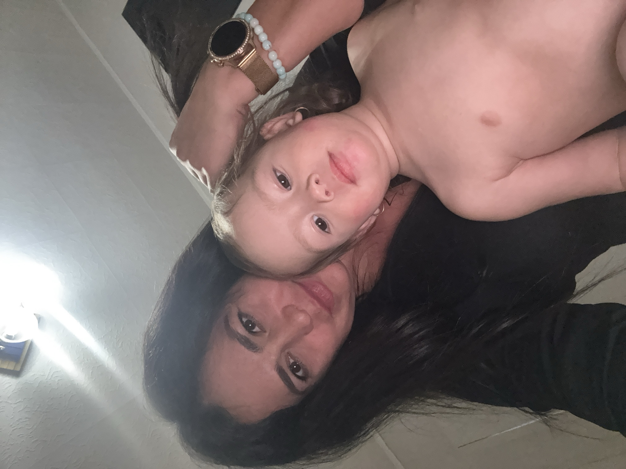 The image, taken in Chaikovsky, Russia, presents an intimate overhead portrait of two individuals, presumably a mother and child, lying down together and looking directly at the camera. The photograph is oriented with a 90-degree clockwise rotation.
In the lower portion of the rotated image, an adult woman is visible, characterized by her long, dark hair and dark eyes. She has a calm and gentle expression, with her head resting on a dark surface. On her right wrist, she wears a rose gold smartwatch with a black, crystal-embellished face and a mesh band, accompanied by a light blue beaded bracelet.
Resting closely beside the woman's head is a young child, likely a toddler, with light brown or blonde hair and light-colored eyes. The child's face shows a curious, slightly smiling expression with rosy cheeks. The child's upper torso is bare, revealing a noticeable light brown birthmark or mole on the left side of their abdomen. A small hoop earring is visible in the child's ear.
The background shows a light-colored, textured ceiling. A bright artificial light source is prominent in the upper left, creating a significant lens flare. A small, framed object is partially visible below this light. The scene captures a moment of close bond and affection in what appears to be an indoor setting. The artificial lighting suggests either an evening shot or an interior space relying on electric illumination. There is no visible text in the image.