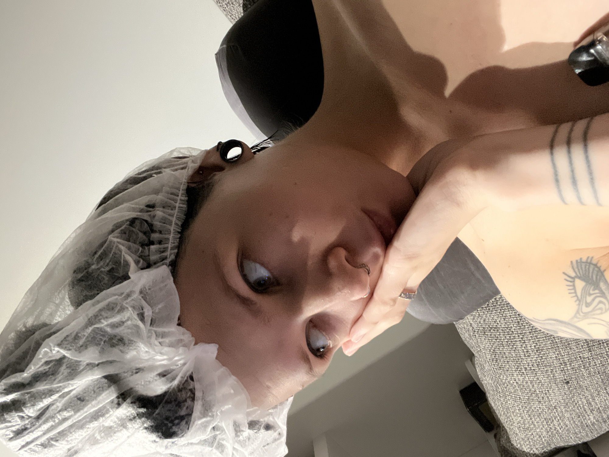 A person with a clear plastic shower cap on their head, is looking upwards, with their hand covering their mouth. They have dark hair, gauged ears, a nose ring and tattoos on their arm and chest. The background includes a gray textured surface and what appears to be a pillow. I do not see anything to suggest the location is Khimki, Russia.
