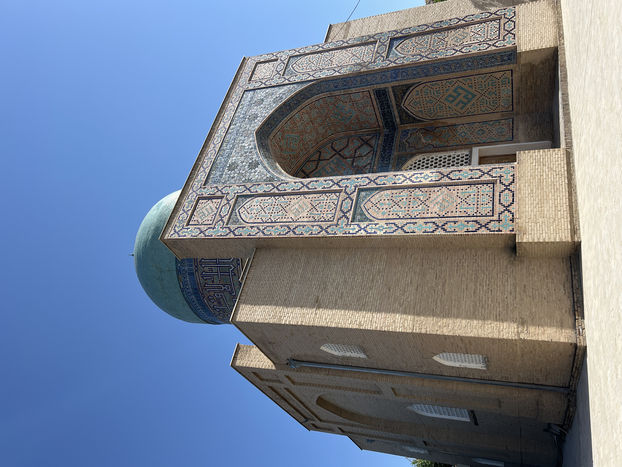 This is a low-angle, eye-level outdoor shot of a building in Samarkand City, Uzbekistan, under a clear blue sky. The structure is primarily made of light brown brick. The main visible facade features a large, ornate archway decorated with intricate blue, turquoise, and pink tilework, forming geometric patterns and Arabic calligraphy. Above the arch, the building continues with more tiled decoration and a large turquoise dome, also adorned with blue tilework. The left side of the building has a flat brick wall with arched windows that have white latticework. The ground in front of the building is paved with light-colored square tiles. There are no people or animals visible in the image. The architectural style suggests a historical religious or governmental building, consistent with the prevalent styles in Samarkand.