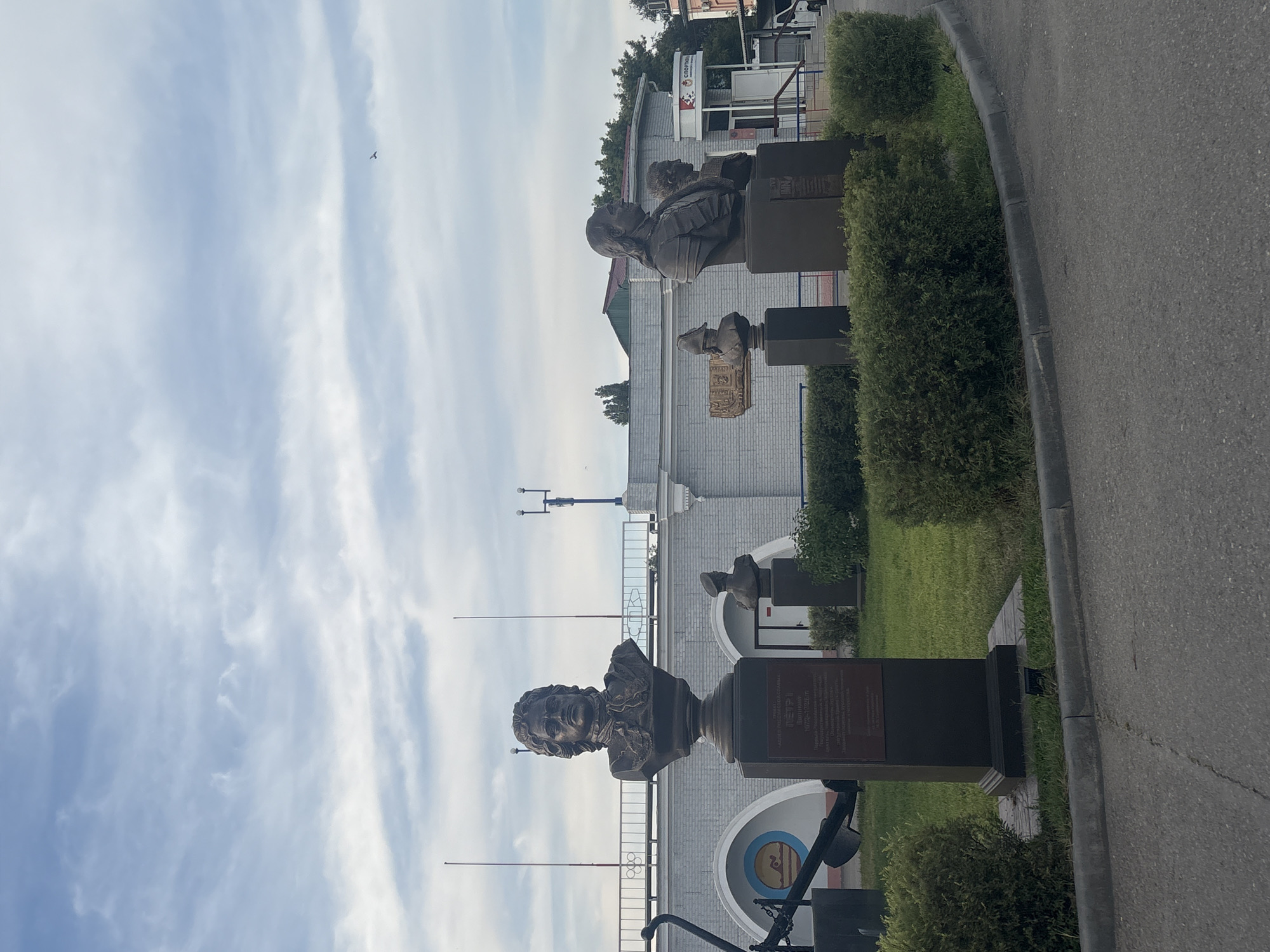The photograph captures a frontal view of a row of busts on pedestals situated on a grassy area in front of a low-rise building. The bust in the foreground, larger than the others, is of a man with curly hair and a neckerchief. A plaque on its dark rectangular pedestal is inscribed with text in Russian, including the name "ПЕТР I" and dates "1672-1725". To the right of this prominent bust are two smaller busts, one of which appears to be a military figure in a cap, and further to the right, a bust of a man with long hair, also on a pedestal. Behind the busts, a white brick building with arched openings and a grey, white, and blue railing is visible. An anchor is partially visible on the left side of the frame. The environment is an outdoor public space with manicured bushes lining the foreground and a paved area with a curb in front of them. The sky above is cloudy with patches of blue. A sign with Cyrillic lettering and an image of a leaping dolphin is visible within one of the building's arched openings. A red and white sign with Cyrillic lettering and an Olympic-style ring symbol is also visible on the building. The scene is static, with no people actively engaged in any actions. The location is identified as Astrakhan, Russia.