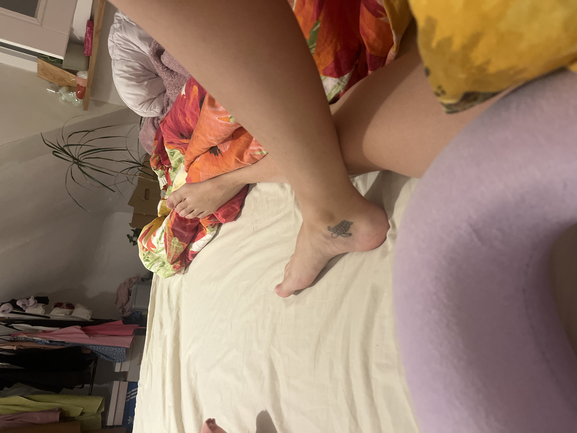 The photograph shows a human subject lying in a bed. The bed has a white sheet and a floral-patterned blanket. A lavender-colored neck pillow is visible. The subject has bare legs and feet, with a turtle tattoo on one ankle. Background includes a shelf with various items, a potted plant, and clothing. The location is an attic room. The city of Saarbrücken, Germany, cannot be determined from the content of this image.