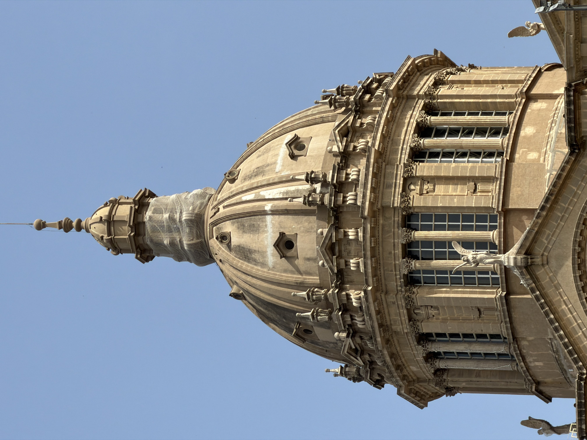 This media file features a close-up, low-angle shot of the exterior of a grand, ornate building, likely a cathedral or a significant historical structure, set against a clear, bright blue sky. The architectural style appears to be Baroque or Neo-Classical, characterized by intricate stonework, columns, arched windows, and decorative gargoyles or sculptures. The dominant subject is a large dome, partially obscured by scaffolding or netting at its apex, suggesting ongoing restoration or maintenance work.
The building's facade is made of weathered stone, displaying a warm, sandy hue. Rows of rectangular windows with multiple panes are visible, framed by classical architectural elements such as pilasters and cornices. Small, decorative sculptures, possibly angels or mythical creatures, are perched on ledges and above the windows. The overall impression is one of historical grandeur and elaborate craftsmanship.
The bright blue sky indicates a daytime scene, likely under fair weather conditions, with no clouds visible. The lighting suggests the sun is high, casting some shadows that highlight the building's three-dimensional features. The absence of people or any signs of daily activity directs the focus entirely on the architectural marvel.
The image is oriented with the building extending upwards, giving a sense of scale and height. The pointed finial at the very top of the dome, crowned with decorative spheres, is a prominent detail. Location cues suggest Barcelona, Spain, known for its rich architectural heritage, which would be consistent with the style of the building. No text is visible in the image.