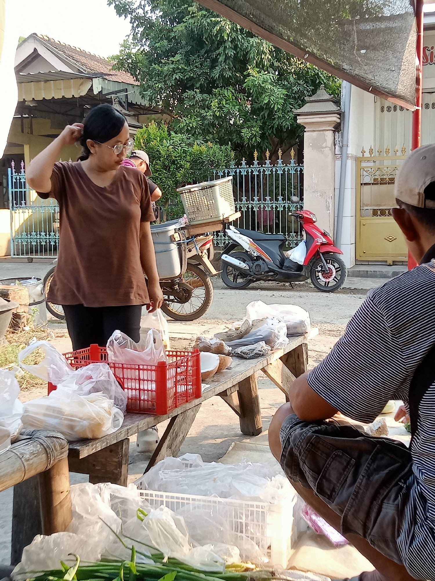 In Simo, Indonesia, an outdoor market scene unfolds. A woman with glasses and dark hair, wearing a brown t-shirt and black pants, stands to the left, looking slightly to the right and touching her hair. In front of her, a wooden table holds several red plastic crates and white plastic bags filled with various items, possibly food products. To the right of the woman, a red motorcycle with a white section and a basket on the back is parked. A man, viewed from the back, wearing a striped t-shirt and dark shorts, is crouched down on the right side of the frame, seemingly attending to items on a lower surface. The background reveals a house with a tiled roof and a yellow gate, as well as lush green trees and a blue decorative fence. The lighting suggests it is daytime, with sunlight casting shadows.