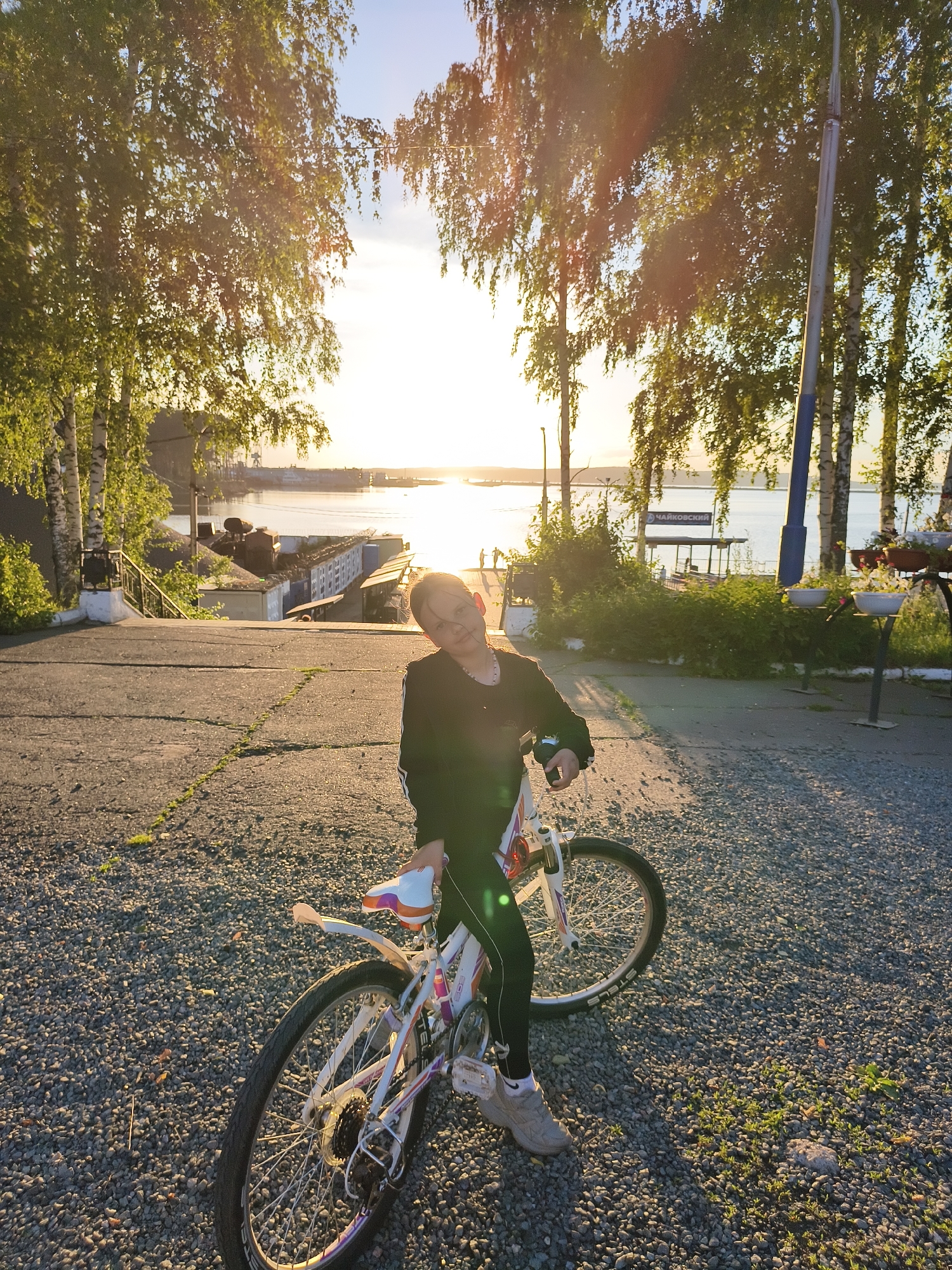 A young person is sitting on a white bicycle with pink and orange accents on a gravel path. The person is wearing black athletic pants and a black long-sleeved shirt. They are looking at the camera with a slight smile and their head is tilted. The bicycle is positioned at an angle, with the rear wheel closer to the viewer. In the background, a wide body of water is visible under a bright, setting sun, which casts a warm glow on the scene. Trees with green leaves line the path and the waterfront, and there are some industrial buildings and structures visible across the water. A lamppost stands to the right of the frame. The path appears to be made of asphalt with patches of gravel and some cracks. The overall scene suggests an outdoor recreational setting in a city, possibly near a river or lake, during the golden hour of sunset.