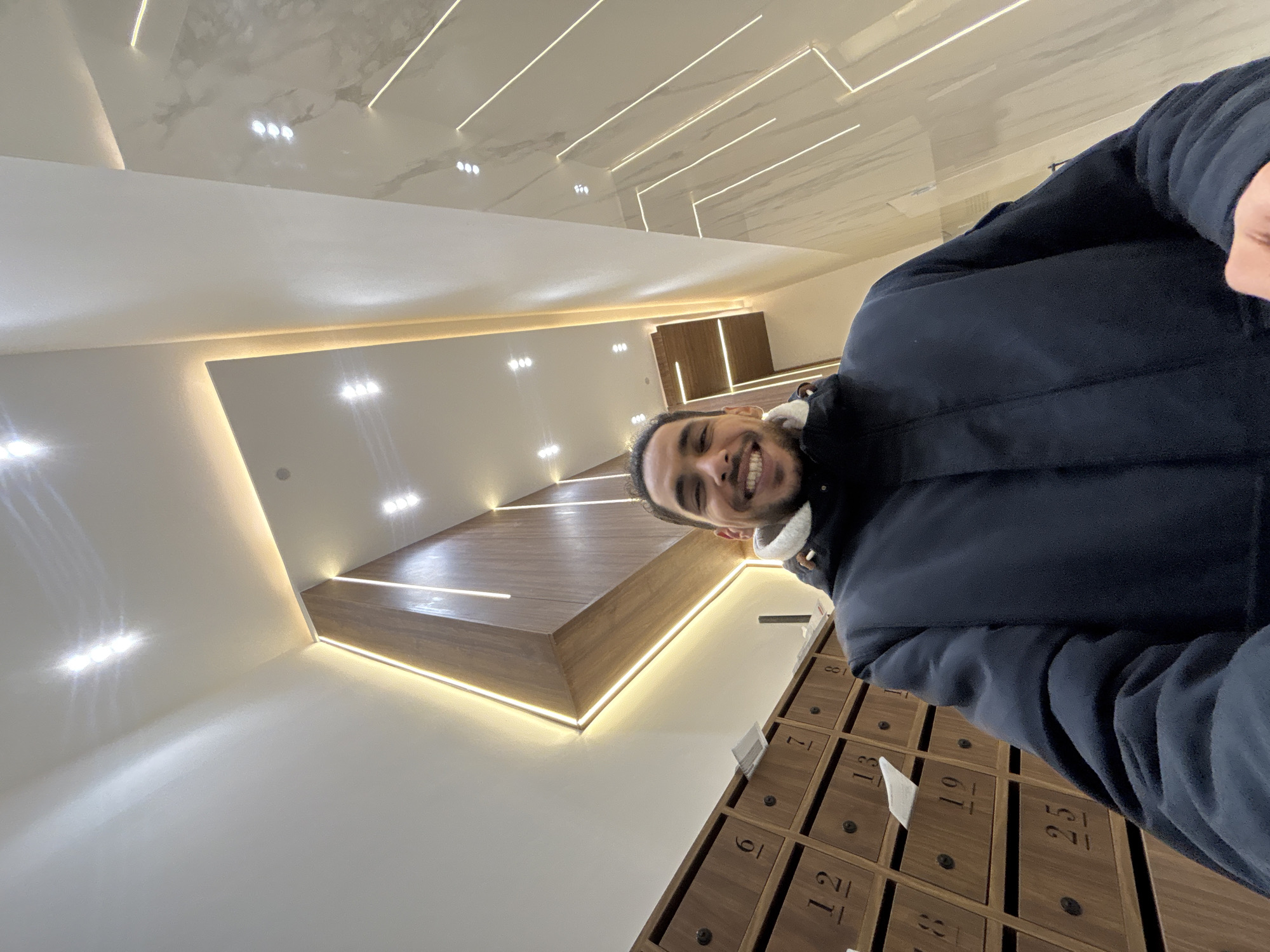An indoor scene, likely a modern residential or commercial building lobby, is captured in Tanger, Morocco. The primary subject is a male individual, presenting with a dark complexion, short dark hair, and a beard, smiling directly at the camera. He is wearing a dark blue hooded jacket with a visible light-colored interior lining at the collar. To the left of the frame, a wall-mounted unit contains numerous wooden mailboxes, each numbered sequentially (e.g., '6', '7', '8', '12', '13', '19', '25'). Some compartments contain visible papers or mail. The upper portion of the scene, encompassing the ceiling and upper walls, is characterized by modern architectural design featuring white surfaces, recessed spotlights, and prominent linear LED strip lighting that delineates geometric patterns and highlights structural elements. The wall on the right side of the frame is covered with light-toned, possibly marble or high-gloss tile, panels that also incorporate vertical and angled linear LED lighting.