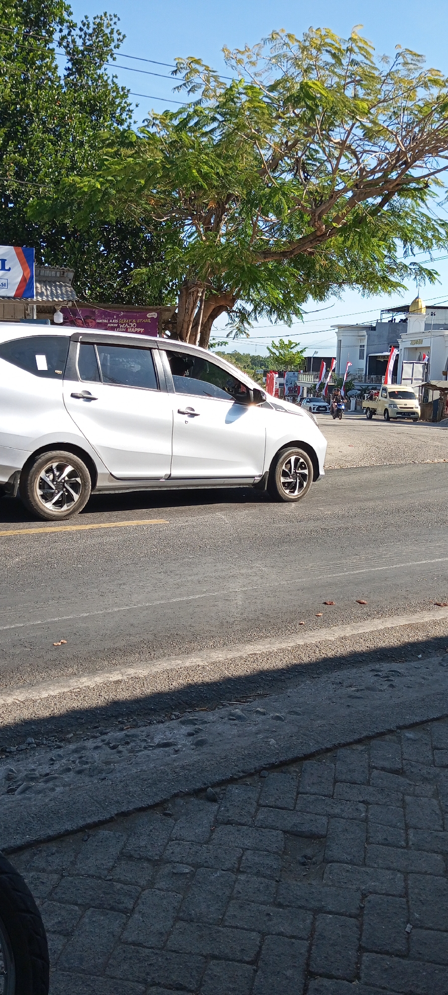 A white car is parked on the side of a road in Pakkanna, Indonesia. The car is a four-door sedan and is parked facing left. The car has tinted windows and a silver trim around the windows. The car's wheels are silver and black. The road is paved with asphalt and has a yellow line painted in the middle. There are trees and buildings in the background. The sky is clear and blue. There are some red and white flags in the background. A yellow truck is parked further down the road. There are a few people on motorcycles in the distance. The scene appears to be a sunny day.
