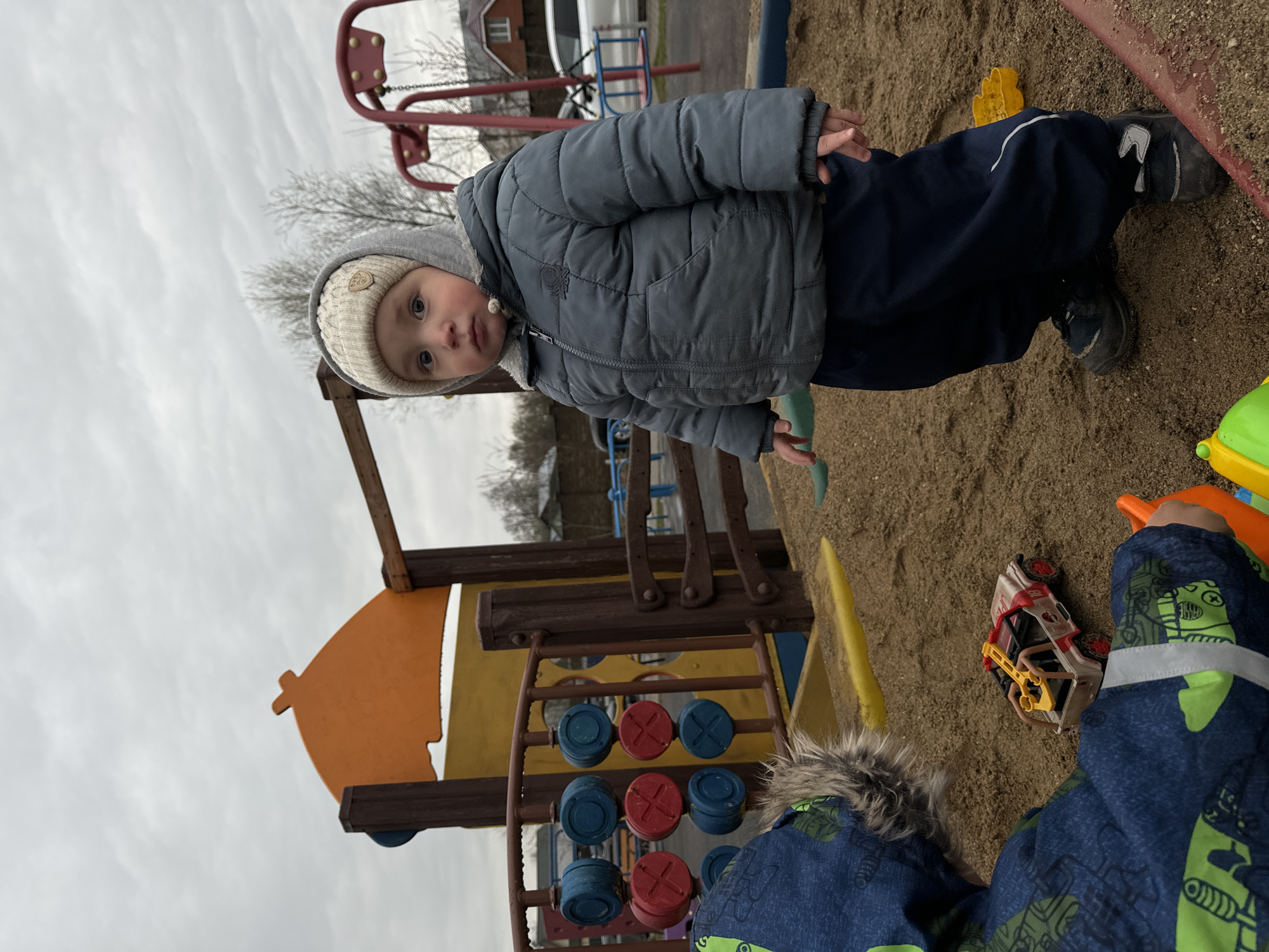 A young child wearing a gray hooded jacket and beige knit hat is standing in a sand-filled playground at a location in Akulovo (Gp Kubinka), Russia. The child is looking directly at the camera. Behind the child is a playground structure with a wooden frame, an orange and yellow sail-shaped element, and a blue and red tic-tac-toe game. In the sand around the child's feet are various toys, including a yellow shovel, a toy car, and other plastic items. Another child in a blue patterned jacket is partially visible in the lower right corner of the frame. The sky is overcast.
