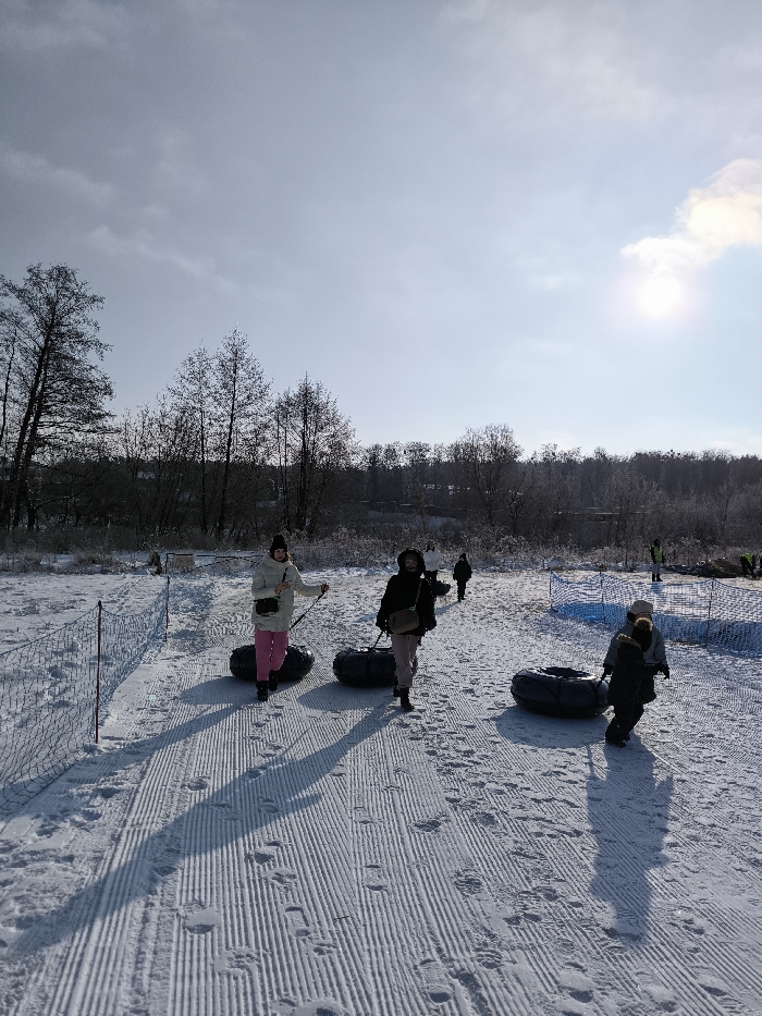 The image depicts a winter scene at a snow tubing area in Prybuzke, Ukraine, on a bright, sunny day. The foreground shows a snow-covered slope with distinct, parallel groomed tracks, indicating a path for walking uphill while pulling snow tubes. Numerous footprints are visible on these tracks.
In the immediate foreground, a total of five individuals are visible, progressing uphill from left to right.
* On the left, a woman wearing a white puffer jacket, pink pants, and a dark beanie is pulling a black snow tube by its rope. She holds a light-colored object in her left hand, possibly a phone or a small bottle.
* To her right, slightly behind, a second woman in a dark jacket, light-colored pants, and a dark beanie is also pulling a black snow tube. Her face is partially obscured by shadow.
* Further to the right and slightly behind, a child in a dark jacket is pulling a third black snow tube. An adult figure, largely obscured, walks behind or alongside this child.
* In the mid-ground, two additional figures are seen walking uphill, one of whom is distinct in a white jacket and dark pants, and another smaller figure behind them.
To the left of the groomed path, a low orange safety net barrier runs along the edge of the snow. Further right, a blue netting also appears to delineate a section of the snow. The background features a dense line of bare deciduous trees and some coniferous trees, suggesting a forest or treeline bordering the recreational area. In the far background, faint structures are visible amidst the trees. The sky is bright blue with scattered white clouds, and the sun, positioned in the upper right quadrant, casts long, prominent shadows from the individuals onto the snow, indicating a low sun angle typical of winter.
