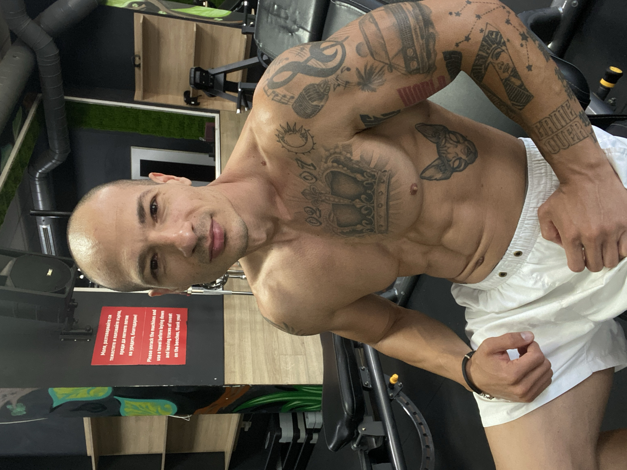 A muscular, bald, shirtless man with extensive tattoos is posing for a photo in a gym. He is wearing white shorts. The gym appears to be modern with equipment visible in the background. A sign in Bulgarian and English is affixed to a black wall to the left of the man, instructing gym users to rack their machines and use a towel. This gym is located in Pleven, Bulgaria, as indicated by the presence of the Bulgarian language on the sign.
