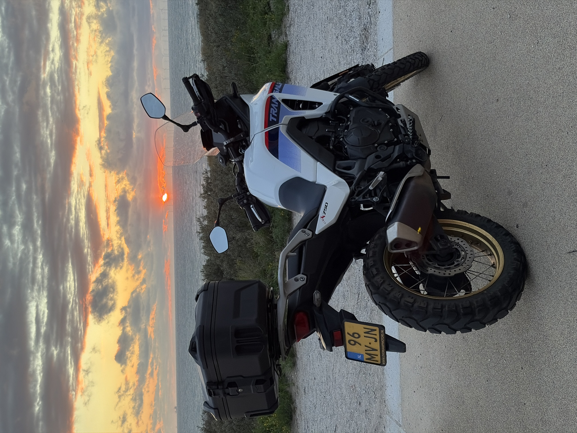 A white and blue adventure motorcycle with a black top box is parked on a paved area near the sea. The motorcycle has a Dutch license plate reading "96 MV-JN". In the background, the sea is visible with a sunset casting a warm glow across the cloudy sky. A line of wind turbines can be seen in the distance on the horizon. The location appears to be a coastal area, possibly a promenade or viewpoint. This scene is consistent with locations in The Hague, Netherlands, that offer coastal views and recreational areas.
