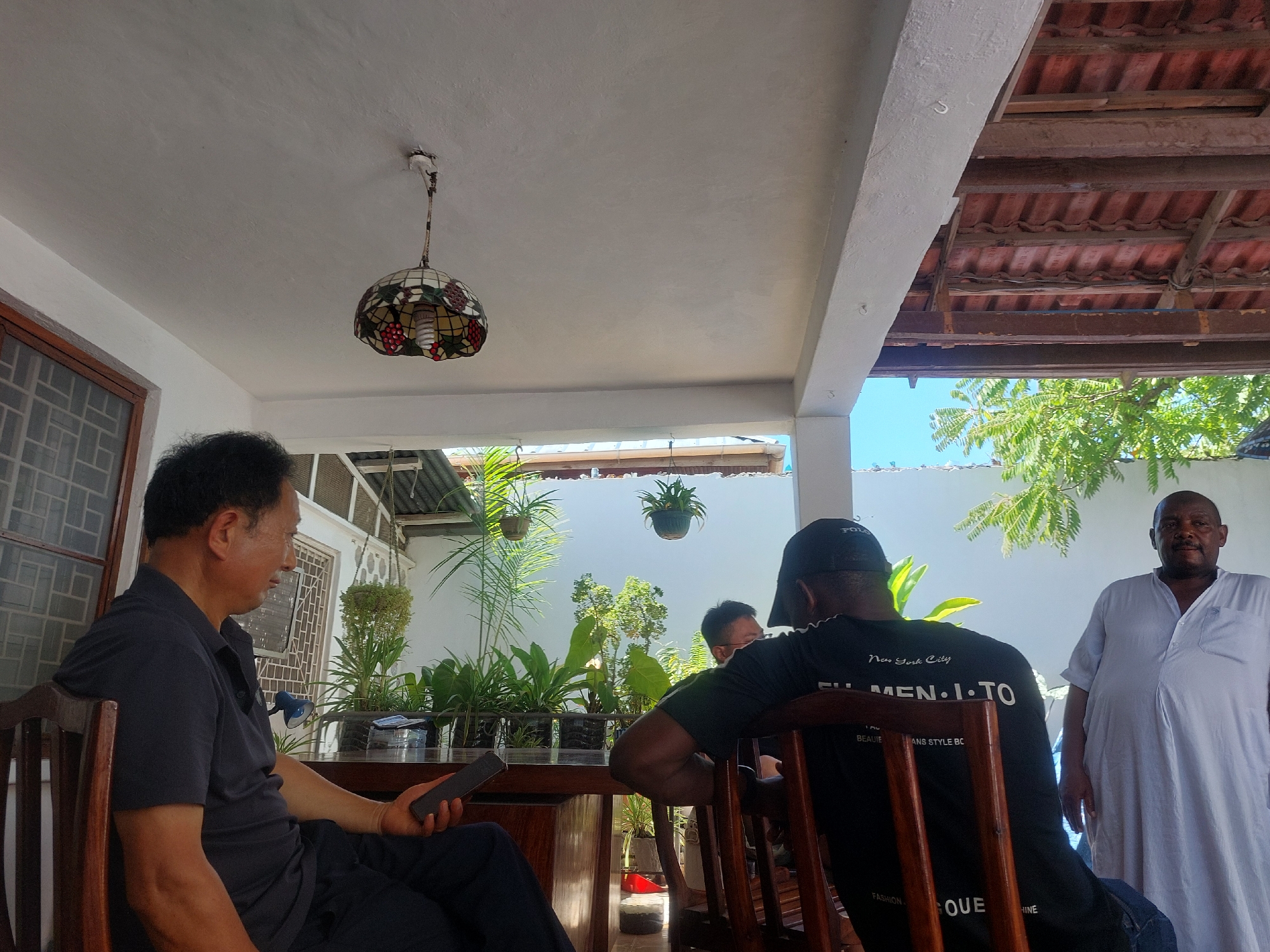 The image captures an outdoor patio scene in Dar es-Salaam, Tanzania. A man in a dark blue polo shirt sits at a wooden table, holding a smartphone. To his right are potted plants on the table. Another man wearing a black t-shirt and a black cap sits with his back to the camera, facing the plants. Further back, a third man in a white robe stands looking towards the camera. Hanging plants are visible in the background. The patio is under a roof with a decorative ceiling lamp. Part of the roof is made with red tiles and wooden beams. Blue sky is visible in the background.