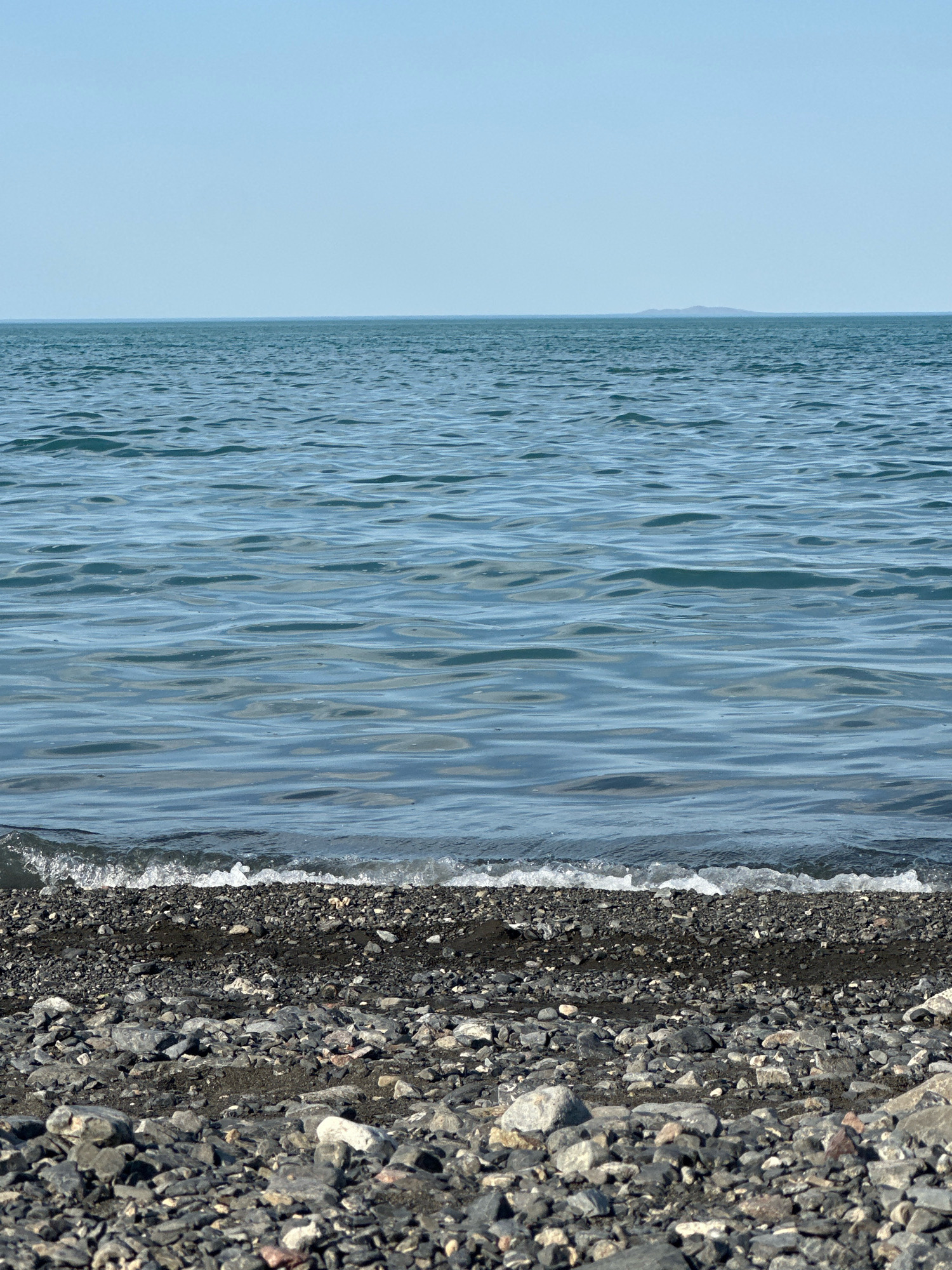 The image depicts a serene natural scene on the shore of Lake Balkhash in the vicinity of Көктұма, Kazakhstan. The foreground is dominated by a pebble beach, comprised of various shades of gray, black, and some lighter-colored stones. A thin line of dark, damp sand is visible between the pebbles and the water's edge. A small wave, with white foam, is gently lapping at the shore. The midground is occupied by the calm, blue waters of Lake Balkhash, reflecting the clear, light blue sky. Subtle ripples are visible on the water's surface, suggesting a light breeze. In the far distance, a faint, low-lying landmass, possibly an island or the distant shore, is barely discernible on the horizon. The overall impression is one of tranquility and natural beauty. There are no people or man-made objects in the frame.