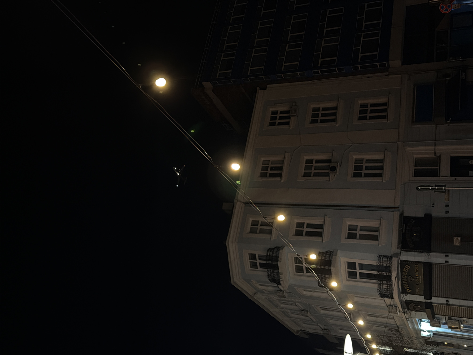 A nighttime exterior shot of a city street, rotated counter-clockwise for aesthetic effect. A pale blue building with multiple windows dominates the right side of the frame. A darker, taller building sits atop the pale blue building. Street lights are strung along wires leading away from the building, creating a line of illumination. The left side of the image is consumed by a dark, unlit space. The location is Unknown, Unknown. A sign reads "Princess London Watches."
