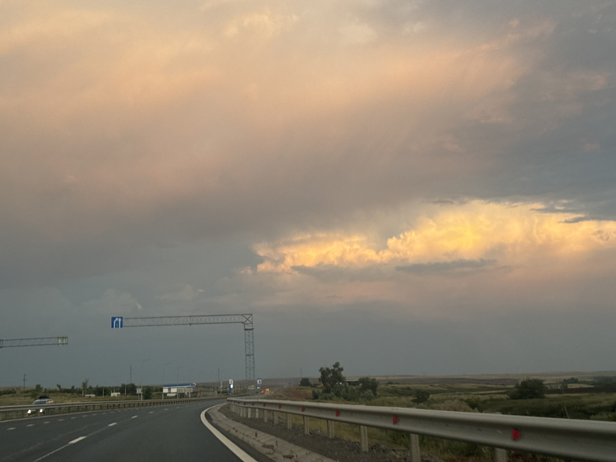 The image depicts a highway curving to the right, with a divided lane. A metal guardrail with red reflectors lines the right side of the road. The sky is filled with dramatic, colorful clouds, with patches of bright orange and yellow light illuminating the upper right quadrant, contrasting with darker gray clouds elsewhere. In the background, a flat, rural landscape with scattered trees and fields is visible. Overhead, a metal gantry with a blue sign displaying two figures is suspended above the road. A few cars are visible on the highway, and a building complex with a white roof can be seen in the distance. The scene appears to be a view from a moving vehicle. There are no explicit landmarks identifying the specific location within the city of Syrt, Russia, but the overall setting suggests a highway passing through a rural area.