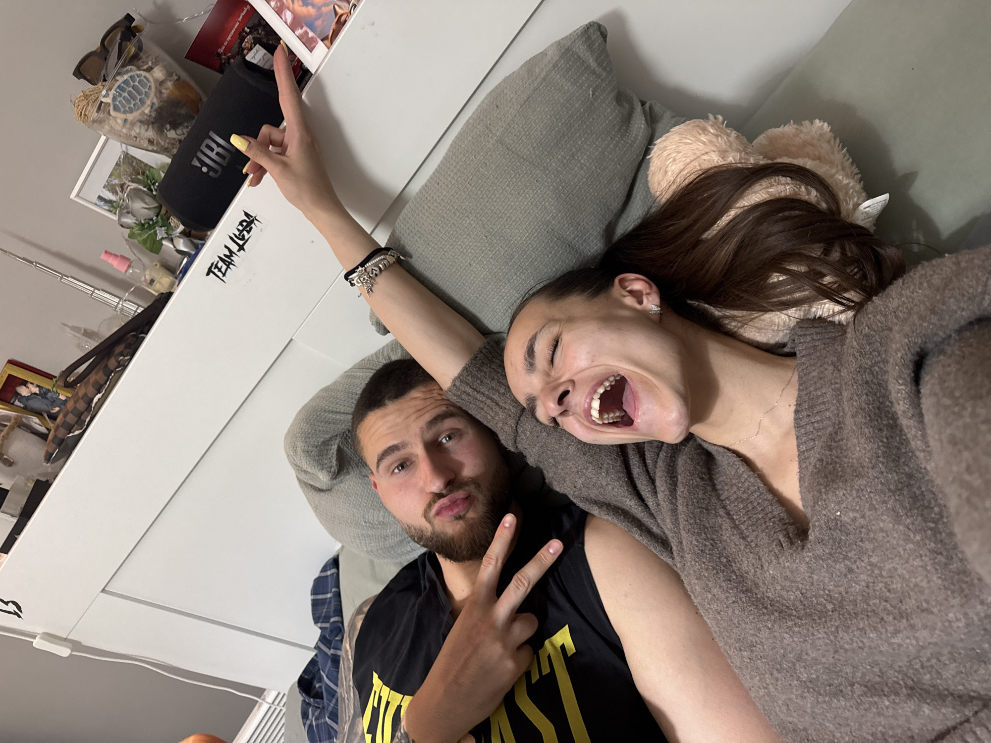A selfie-style photo captures two young adults, a man and a woman, lying down indoors, likely in a bedroom in Sofia, Bulgaria. The image is taken from a slightly elevated, diagonal perspective.
The man, on the left, has a beard and short dark hair. He is wearing a black sleeveless top with a yellow graphic (partially visible, possibly "EVERLAST") over a plaid shirt, and is making a peace sign with his right hand while looking directly at the camera with a slightly pursed-lip expression.
The woman, on the right, has long dark hair and is wearing a brown knitted sweater and a delicate silver necklace. She is looking upwards and outwards with a wide-open mouth, suggesting a playful shout, laugh, or exaggerated expression. Her right arm is raised, and she is making a peace sign with her fingers pointing towards a shelf behind her head. She also has bright yellow nail polish and a multi-charm bracelet on her wrist.
They appear to be lying on pillows, one grey and textured, the other light brown and plush, resembling a furry cushion or stuffed animal. Behind them is a white headboard or shelf unit. On this shelf, various personal items are visible: a black JBL portable speaker, sunglasses, a clear jar containing shells and a blue turtle ornament, a small pink spray bottle, and several small framed photographs. Black, stylized text reading "TEAM JUDA" is written on the white surface of the shelf.
The overall mood of the photo is casual, joyful, and relaxed, suggesting a moment of fun between the two individuals. The time of day is not discernible, as the setting appears to be indoors with artificial or ambient lighting.