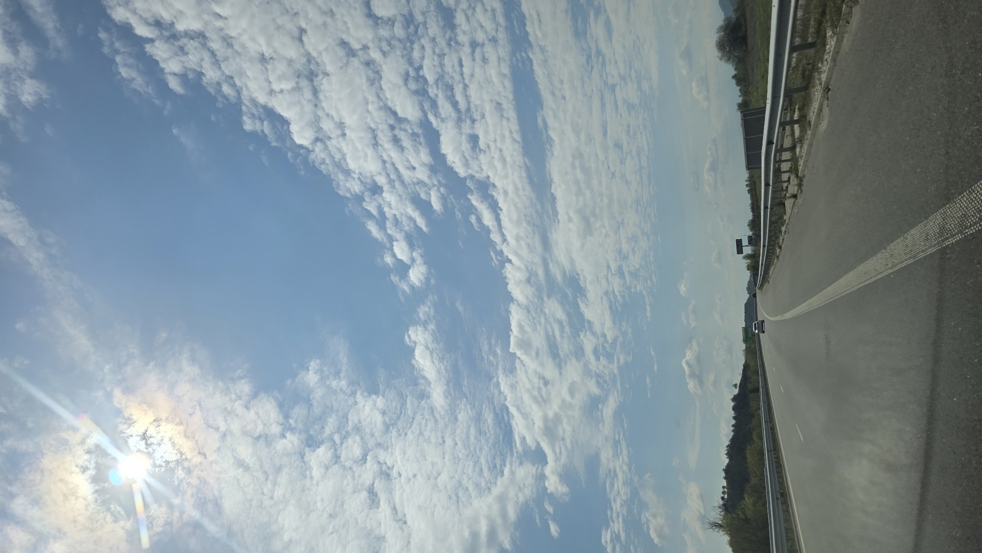 The image shows a highway with a clear blue sky filled with scattered white clouds. The sun is visible in the lower left corner, emitting bright rays. The highway has a shoulder with a metal guardrail. A single white line divides the lanes. In the distance, green trees line the horizon, and a vehicle is visible on the road. This scene is likely situated in the city of Valkovo, Bulgaria.