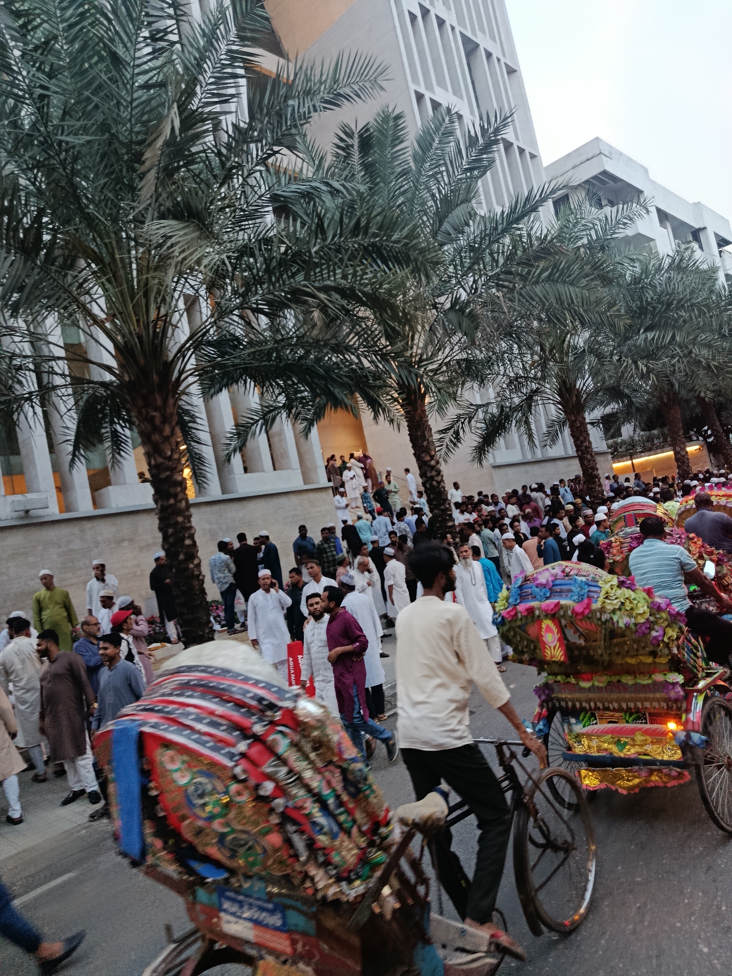 The image depicts a crowded street scene in Dhaka, Bangladesh, likely during a religious or cultural event. A large group of men, many wearing traditional Islamic attire such as thobes and caps, are gathered near a modern building with a distinctive facade. Several date palm trees line the street, providing shade and adding to the atmosphere. Rickshaws, adorned with colorful decorations, are visible in the foreground and moving through the crowd. A rickshaw puller, dressed in a light-colored shirt and dark pants, is pedaling his vehicle. The overall environment suggests a festive occasion in an urban setting.