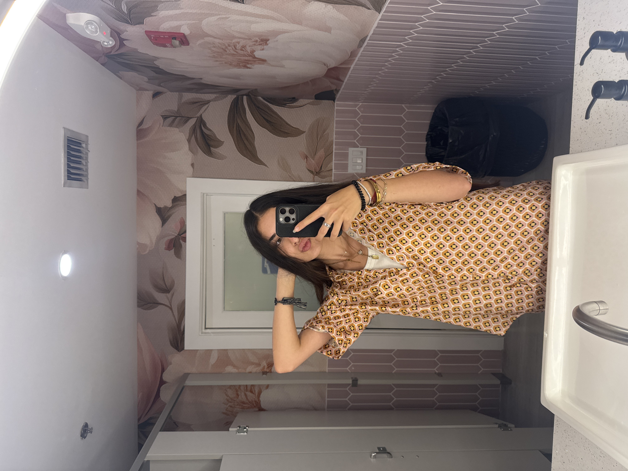 A young woman is taking a mirror selfie in a bathroom. She is wearing a pink and orange patterned shirt over a white tank top, and has her hands behind her head. She is wearing several bracelets and necklaces. The bathroom has floral wallpaper, pink tiled walls, and a white sink with a chrome faucet. There is a black trash can in the corner. The image was taken at an unspecified location in Boca Raton, United States of America.