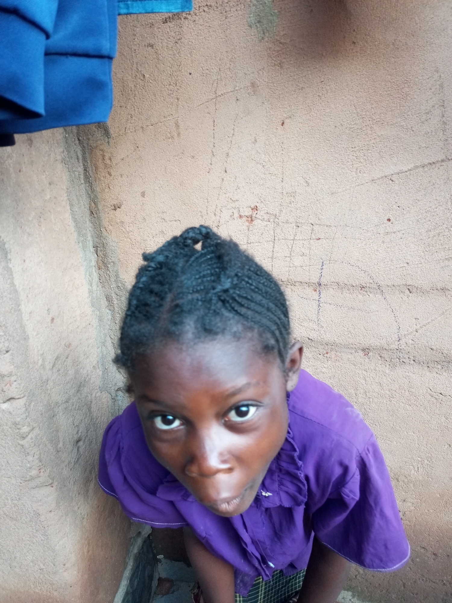 A young Black female is shown from a high-angle perspective, looking up into the camera. She has dark skin and dark, braided hair pulled back into a bun on top of her head. She is wearing a bright purple collared shirt with ruffles around the neckline. A portion of a plaid garment, possibly a skirt or trousers, is visible at the bottom of the frame. The background consists of a textured, light brown wall with some faint markings and a small section of a blue garment to the upper left. The environment suggests an outdoor urban setting, possibly in Gombe, Nigeria, with the rough wall indicating a building exterior. The lighting is somewhat diffused, and there are no other individuals or significant objects in the immediate vicinity. The overall impression is a candid portrait.
