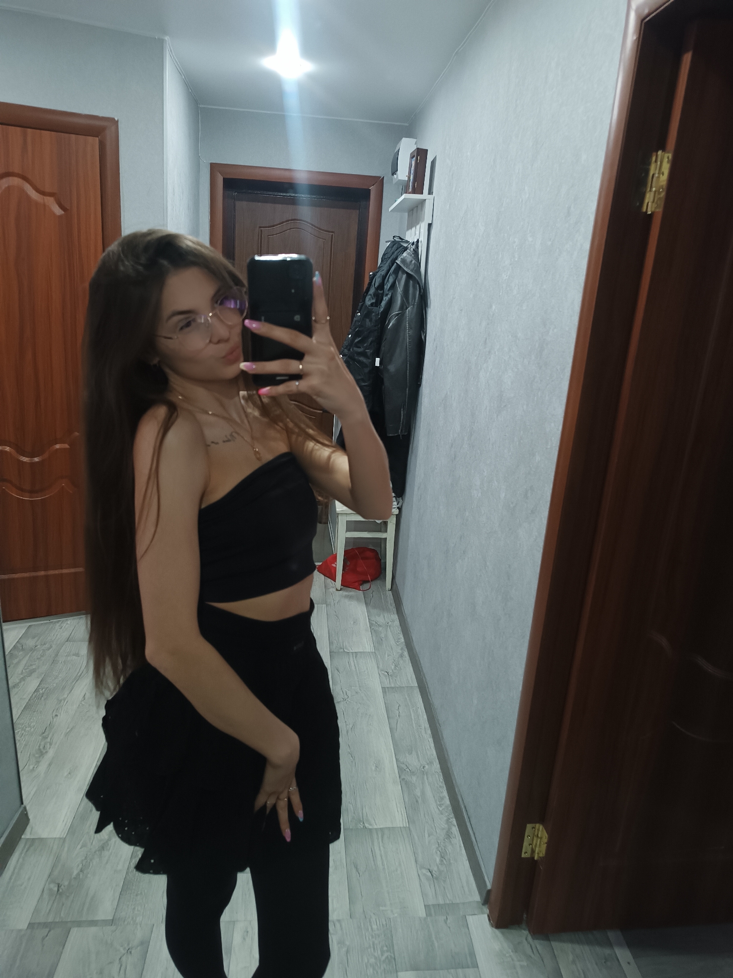 A young woman is taking a selfie in what appears to be the hallway of an apartment or house. The location is Unknown, Unknown.
The woman has long brown hair, wears glasses, and is dressed in a black tube top and a black mini-skirt over black leggings or tights. She is holding a phone in her right hand to take the picture.
The background consists of walls with light grey patterned wallpaper. There are two closed wooden doors, one on the left and one on the right, and another door slightly ajar in the background. A white shelf with items on it and a black jacket hanging on it are also visible. The floor has a light grey wood pattern.