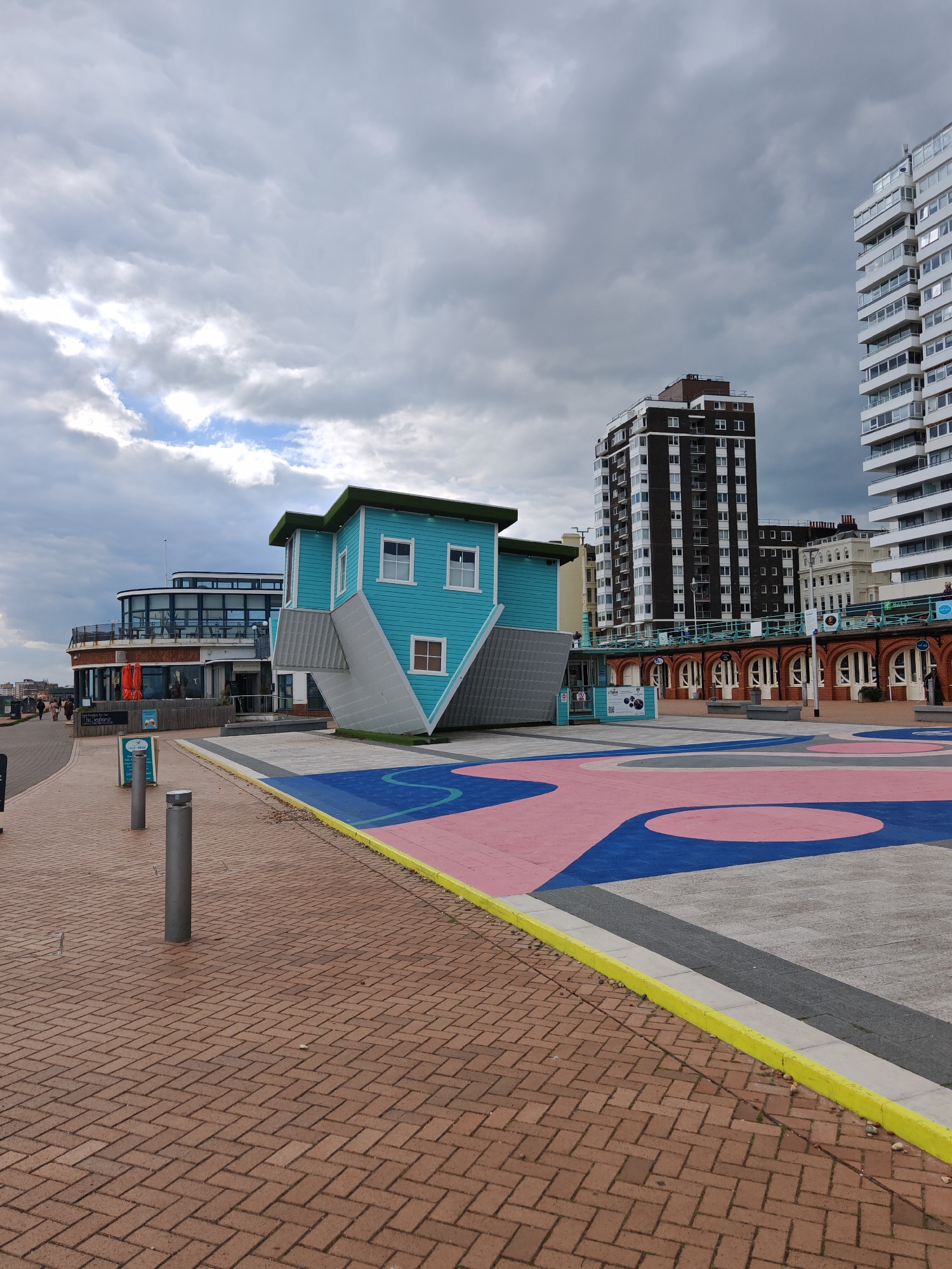 The image shows a structure resembling an upside-down house in a public space, likely a tourist attraction. The house is painted blue with white trim and has a grey roof. It is positioned on a painted concrete area with blue, pink, and grey designs. In the background are various buildings, including a modern building with a curved glass facade and taller, block-style buildings. The foreground includes a brick-paved area with metal bollards. The sky is overcast with heavy clouds. It is not possible to confirm the location in the city of Reigate and Banstead, United Kingdom from the image alone.