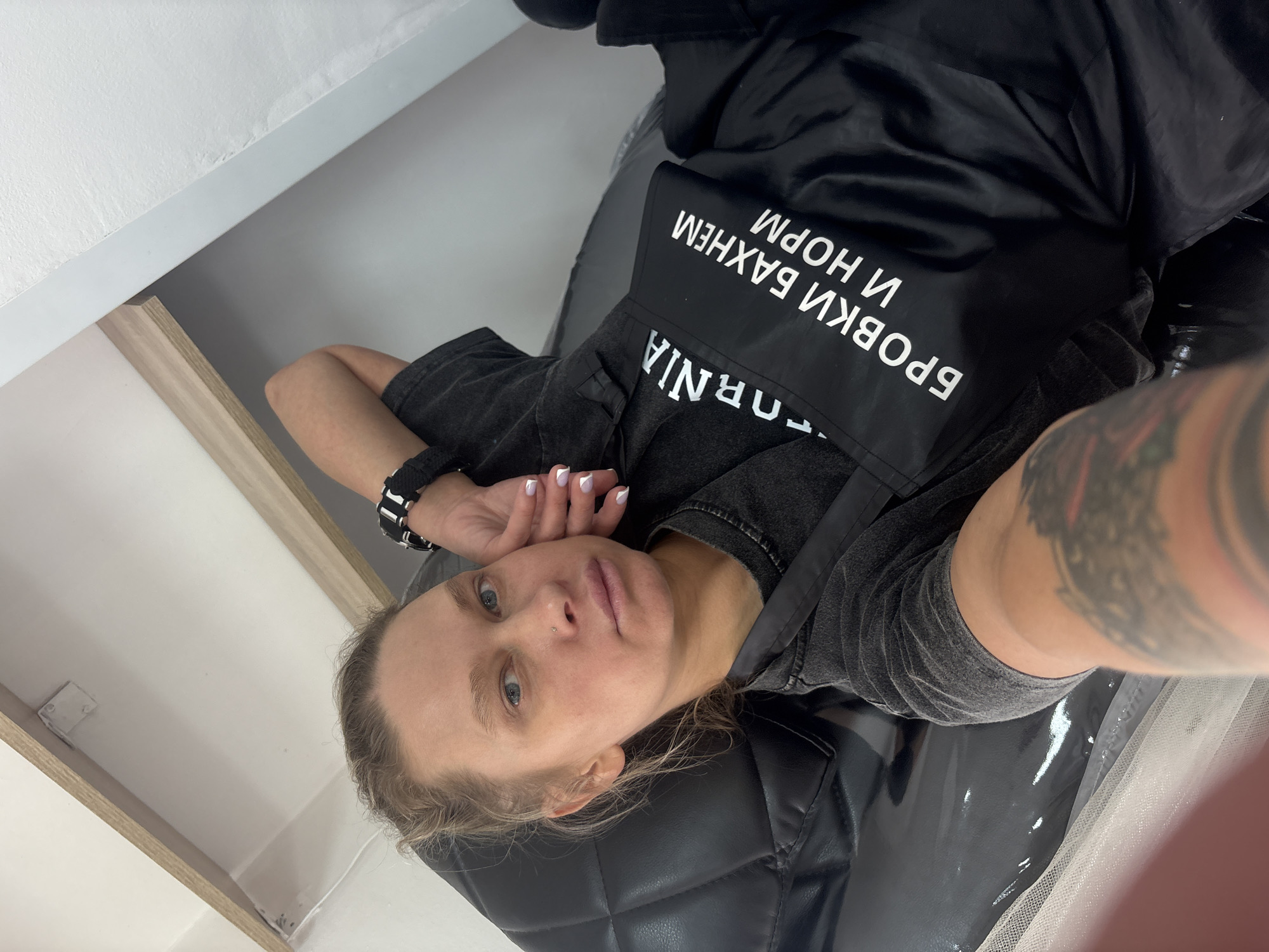 A woman with fair skin, blond hair, and blue eyes is lying on a black quilted surface. She is wearing a gray t-shirt and a black smock with white lettering in Russian that includes the words "BROVKI BAXHEM I NORM." She has a watch on her left wrist and a tattoo on her right forearm. Her left hand is near her face, displaying a fresh French manicure. The setting appears to be inside a building, with white walls and trim visible in the background. The location is likely Perm, Russia, due to the Russian lettering on the smock.