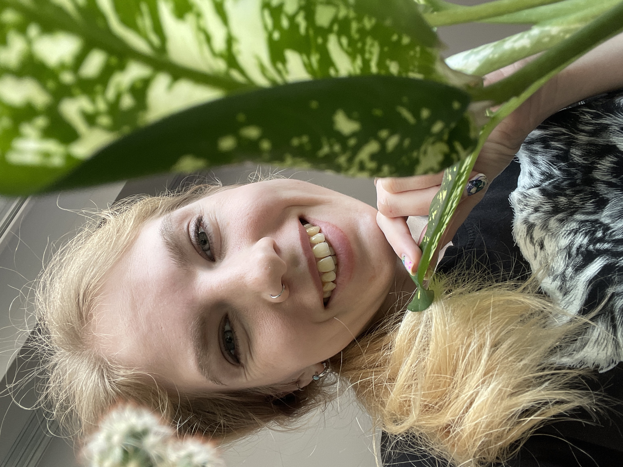 A blonde woman is holding a green and white leafy plant in front of her face. She has a nose ring and is smiling. Her hair is partially visible. In the background, a black and white patterned object is visible. Part of a cactus is on the lower left. The location is not identifiable from the provided image.