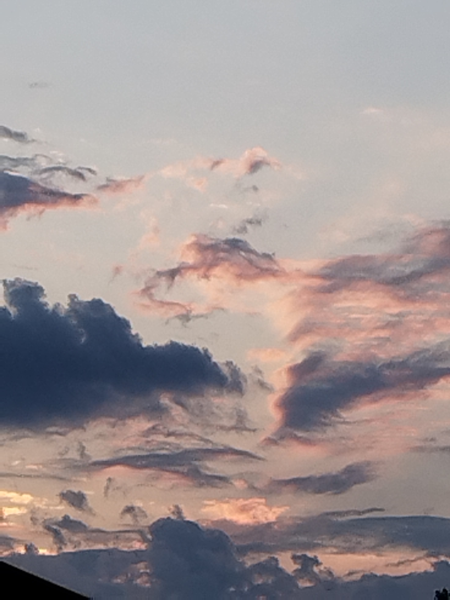 The image shows a sky filled with clouds, some of which are illuminated with pink and orange hues, suggesting it is either sunrise or sunset. Darker, denser clouds are also present, creating a contrast in the sky. The bottom portion of the image shows the silhouette of a dark roofline, indicating an urban or suburban environment. Without further visual cues or contextual information, the specific location within Bucharest, Romania, cannot be determined.
