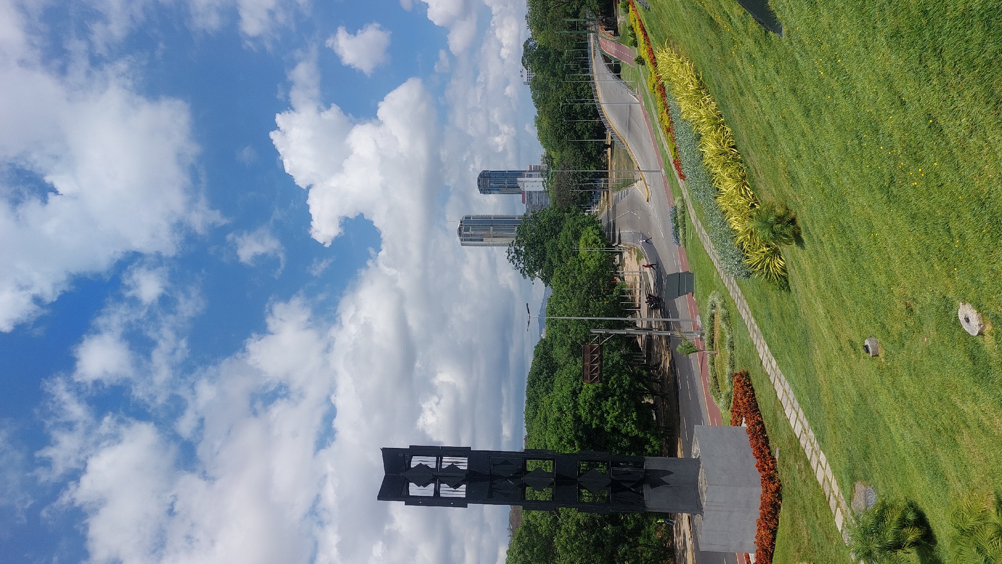 The image shows an upward-angled view of Caracas, Venezuela, featuring a park landscape, cityscape, and sky. A tall, black monument stands in the foreground. A multi-lane road lined with greenery and trees separates the monument from a large grassy area with a stone pathway. Two tall buildings are visible in the mid-ground, set against a partly cloudy sky. People walk along the road and sidewalk.