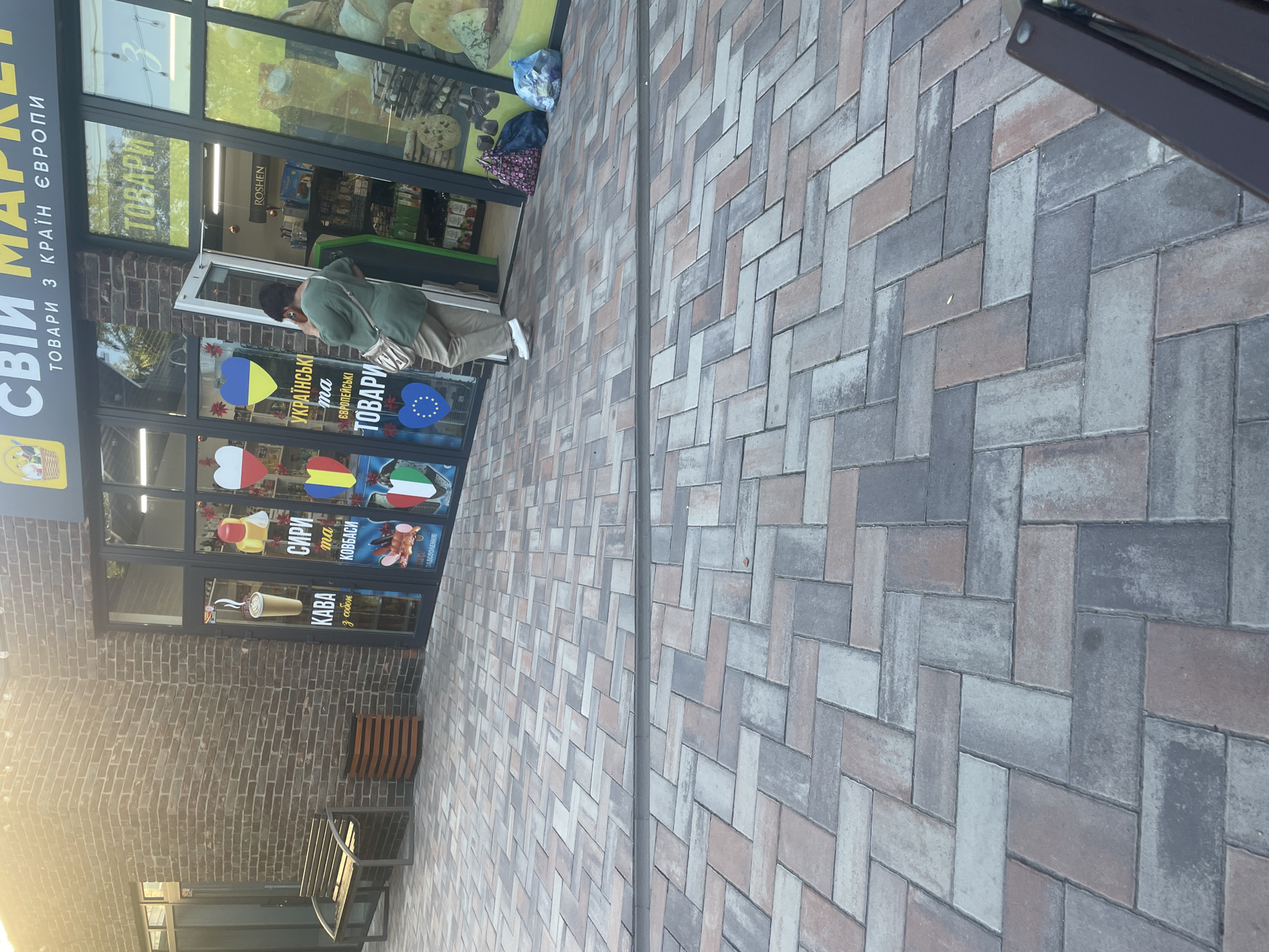 The image shows an outdoor scene in front of a market. The market has a brick facade and large glass windows displaying products. Above the windows, the market's name "СВІЙ MARKET" (SVOY MARKET) is visible in yellow letters, along with the tagline "ТОВАРИ З КРАЇН ЄВРОПИ" (PRODUCTS FROM EUROPEAN COUNTRIES). The entrance to the market is open, and a woman in a green top and beige pants is standing just inside, holding a phone to her ear. Inside the market, shelves are stocked with various goods, including food items and confectioneries. Outside the market, to the left, there is a wooden bench and a dark-colored trash receptacle. The ground in front of the market is paved with interlocking stones in various shades of gray, brown, and beige, arranged in a herringbone pattern. There are two bags, one blue and one with a pink and purple pattern, placed on the ground near the entrance. The overall environment suggests a commercial area, possibly in a town or city.