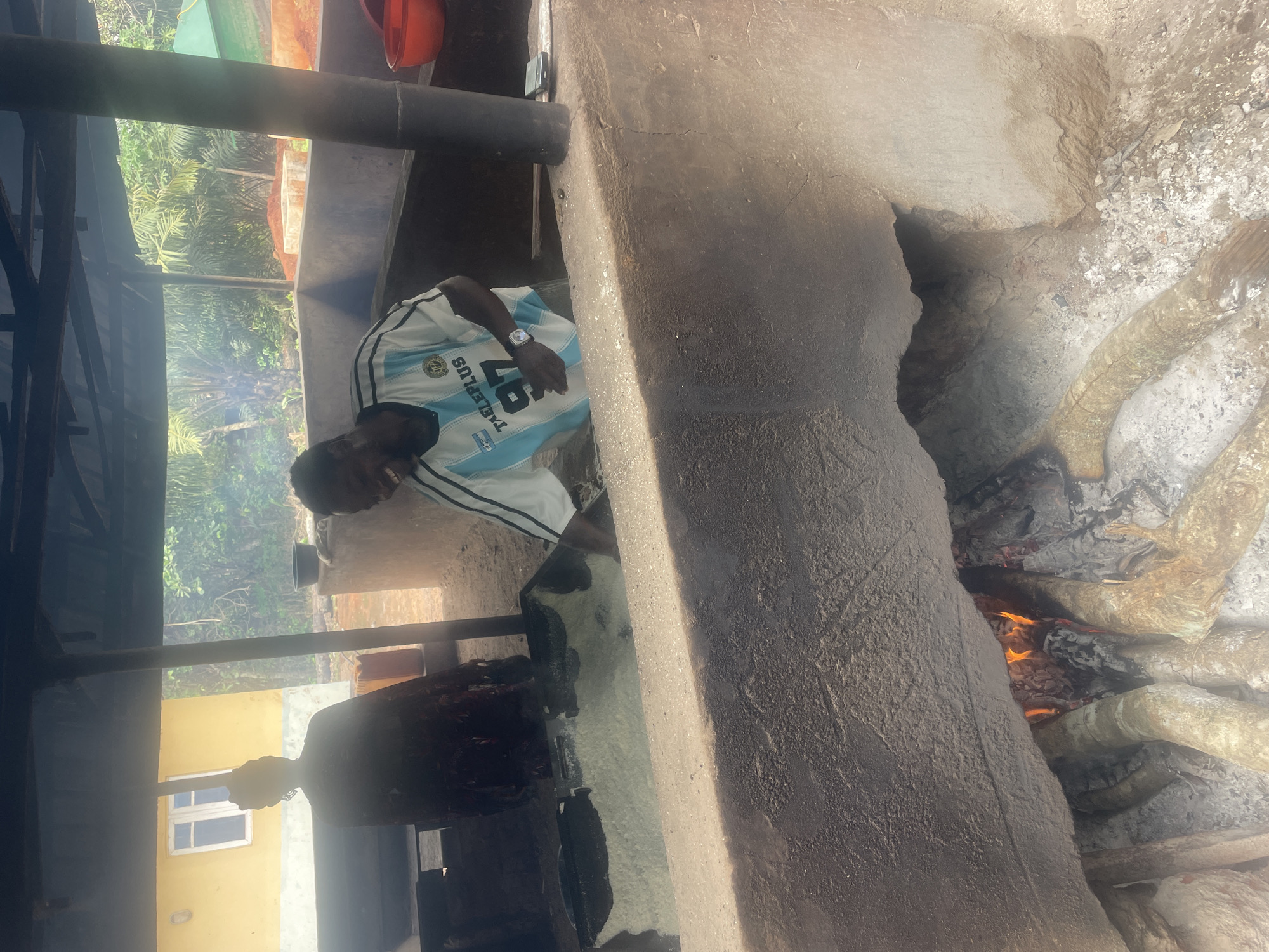 A man wearing a white and light blue soccer jersey with the number "97" on it stands next to a large outdoor stove in Eke, Nigeria. The stove is constructed of concrete and appears to have a fire burning inside. The man seems to be monitoring or interacting with something inside the stove. Overhead is a simple metal structure, possibly a shelter. In the background, a yellow building with a white-framed window is visible. The surrounding environment includes trees and greenery, suggesting an outdoor location.